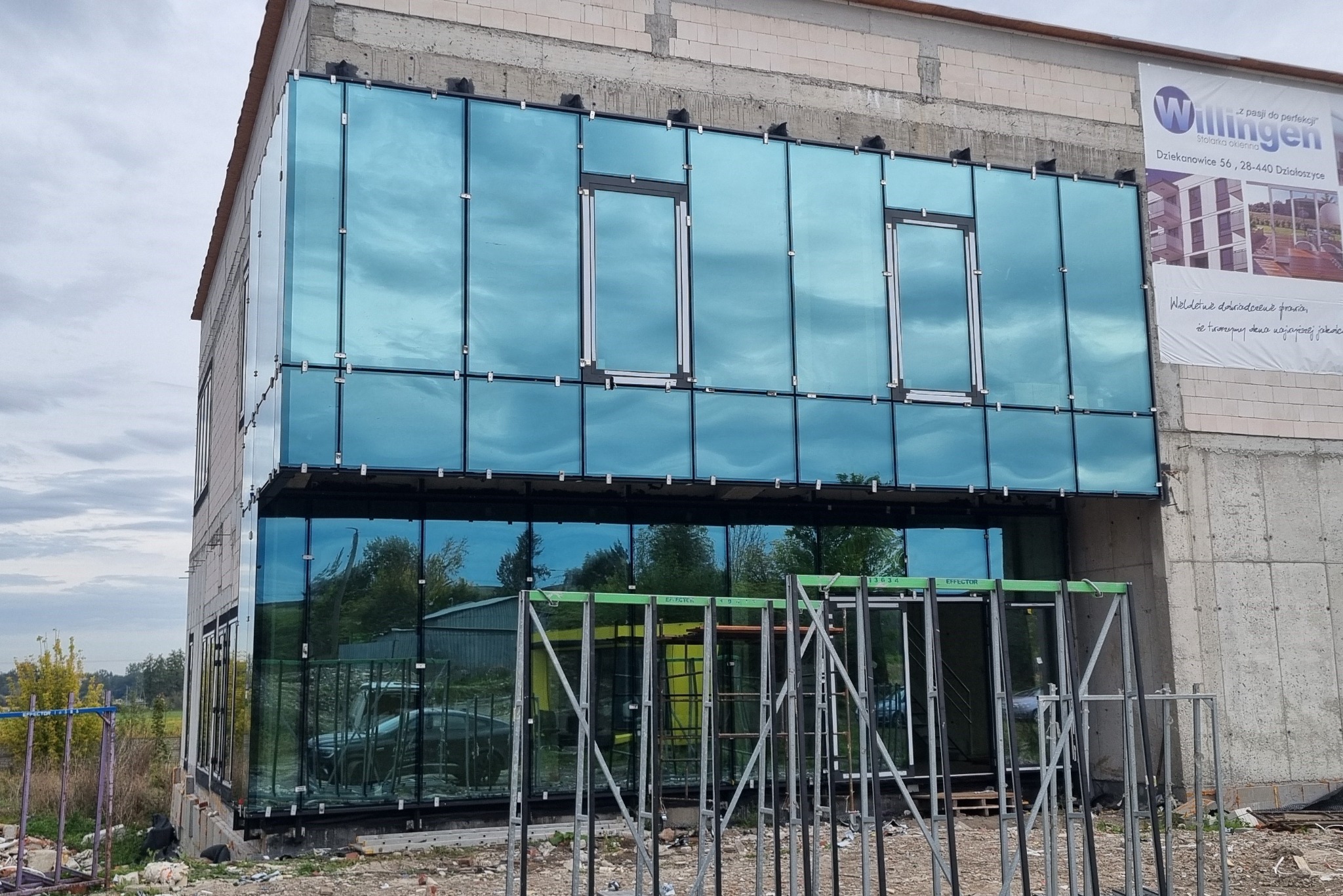 Nowoczesny budynek biurowy w trakcie montażu szklanej fasady z niebieską poświatą, widoczne rusztowania i baner reklamowy firmy.