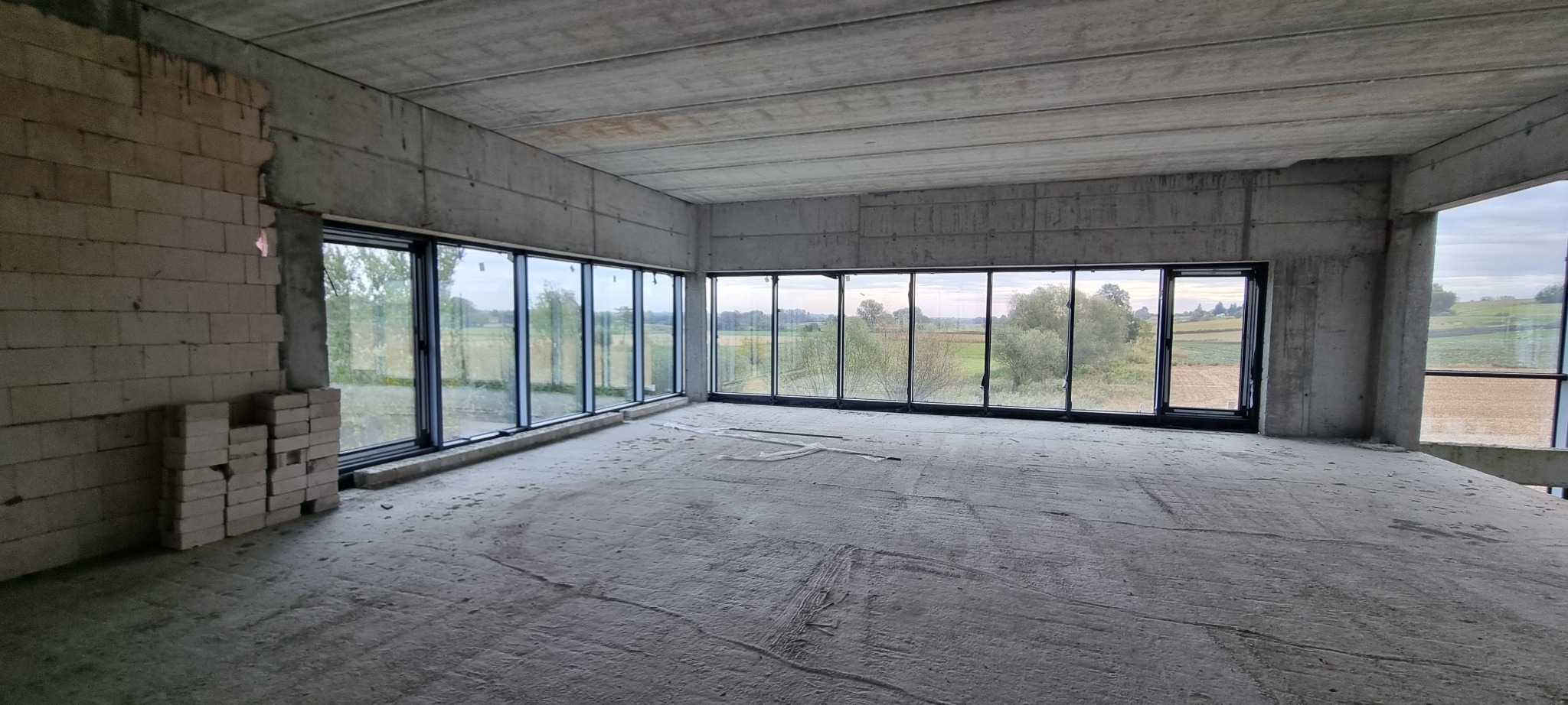 Surowy stan wnętrza z dużymi oknami w czarnych ramach, widok na okolicę Działoszyc, ściany w stanie deweloperskim.