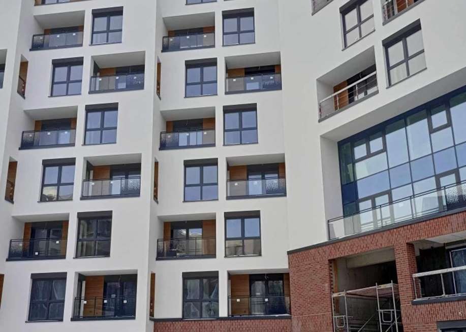 Nowoczesny budynek mieszkalny z ciemnymi oknami i balkonami z przezroczystymi balustradami, fasada w odcieniach bieli i szarości, częściowo wykończona cegłą.