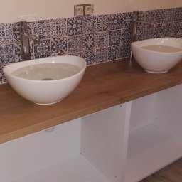 FIRMA REMONTOWO-BUDOWLANA BERT-HOUSE ROBERT KUBACKI - Dwie białe, owalne umywalki nablatowe z chromowanymi bateriami na drewnianym blacie. W tle dekoracyjne, wzorzyste płytki i białe ściany. Widoczne gniazdka elektryczne.