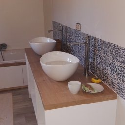 FIRMA REMONTOWO-BUDOWLANA BERT-HOUSE ROBERT KUBACKI - Wykończona łazienka z dwiema owalnymi umywalkami na drewnianym blacie, dekoracyjnymi płytkami i wanną w tle. Widoczne narzędzia glazurnicze.
