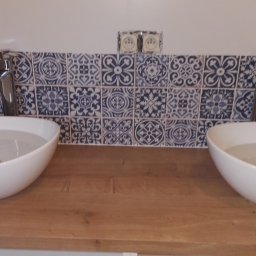 FIRMA REMONTOWO-BUDOWLANA BERT-HOUSE ROBERT KUBACKI - Dwie białe, owalne umywalki nablatowe z chromowanymi bateriami na drewnianym blacie. Nad nimi dekoracyjne, niebiesko-białe płytki ceramiczne i gniazdka elektryczne.