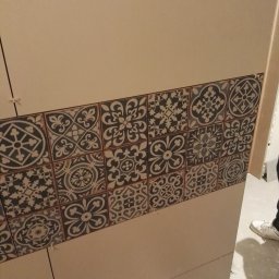 FIRMA REMONTOWO-BUDOWLANA BERT-HOUSE ROBERT KUBACKI - Ściana wyłożona beżowymi płytkami z dekoracyjnym pasem płytek w stylu vintage, z widocznymi krzyżykami dystansowymi, częściowo widoczna osoba w tle.