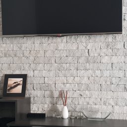 FIRMA REMONTOWO-BUDOWLANA BERT-HOUSE ROBERT KUBACKI - Ściana z dekoracyjnej, szarej cegły z telewizorem LG. Na komodzie ramki ze zdjęciami, świece i dekoracje. Minimalistyczny wystrój wnętrza.