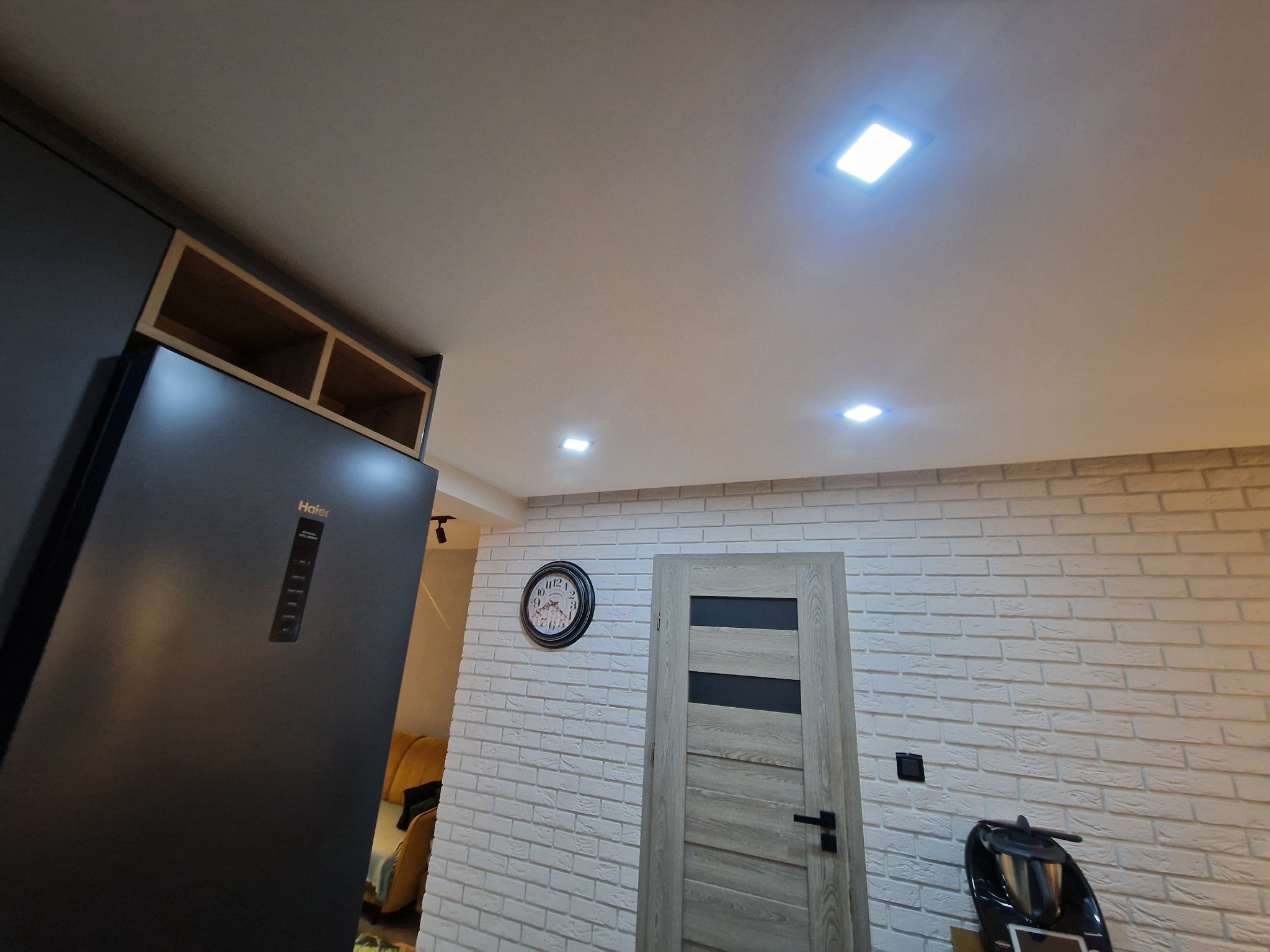 Nowoczesna kuchnia z białą cegłą na ścianie, czarną lodówką Haier i drewnianymi akcentami. Widoczny zegar i drzwi z przeszkleniami. Oświetlenie sufitowe.