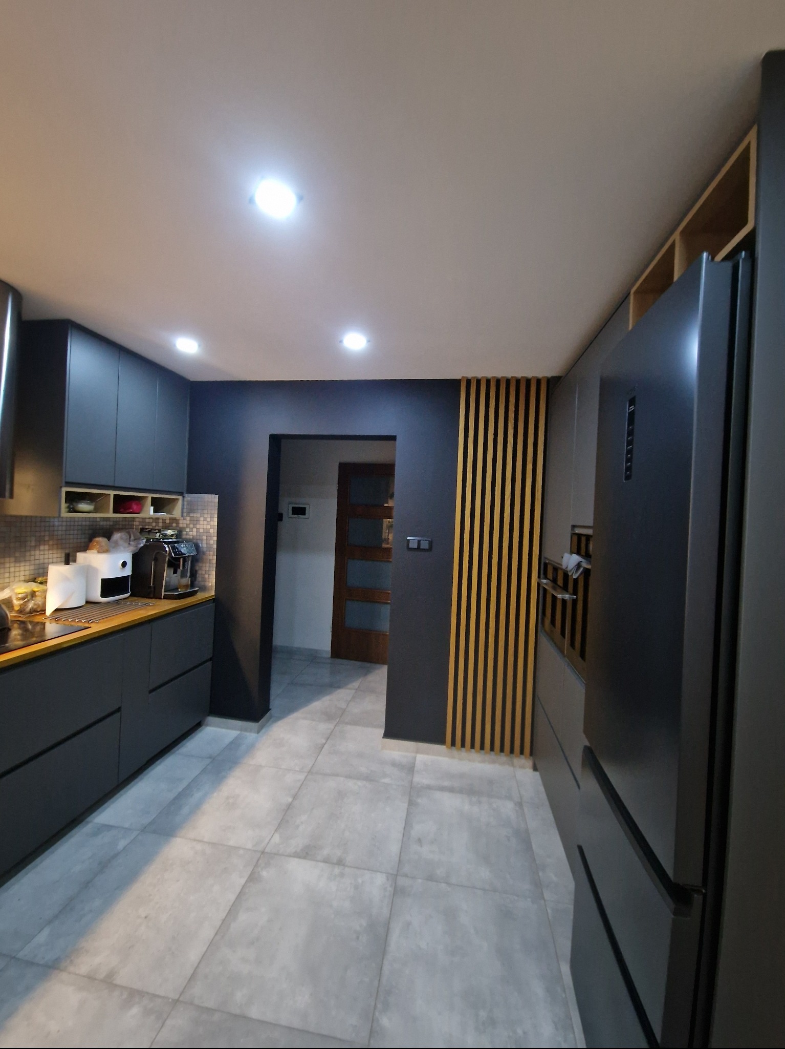 Nowoczesna kuchnia w odcieniach szarości z drewnianymi akcentami. Widoczne szafki, sprzęt AGD i płytki podłogowe. Oświetlenie sufitowe.