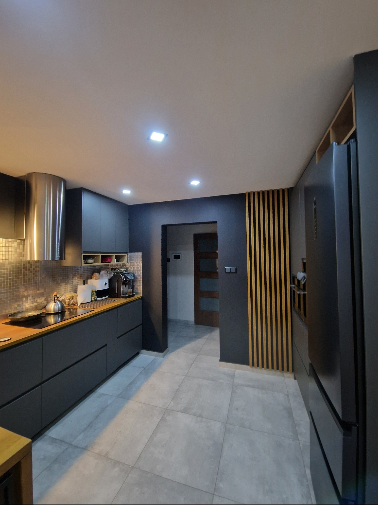 Nowoczesna kuchnia w odcieniach szarości z drewnianymi akcentami. Widoczny fragment blatu, mozaika na ścianie i pionowe drewniane listwy dekoracyjne.