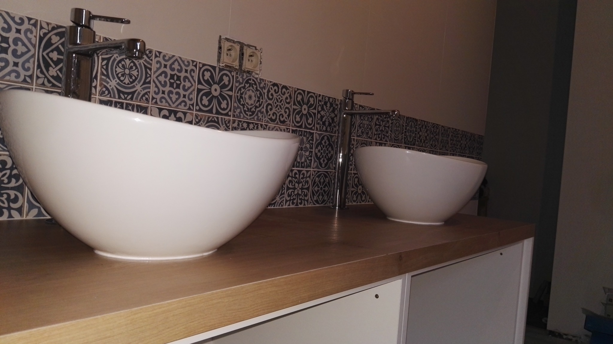 Dwie białe, owalne umywalki nablatowe z chromowanymi bateriami na drewnianym blacie, z dekoracyjnymi, ciemnymi płytkami w tle. Widoczne gniazdka elektryczne.