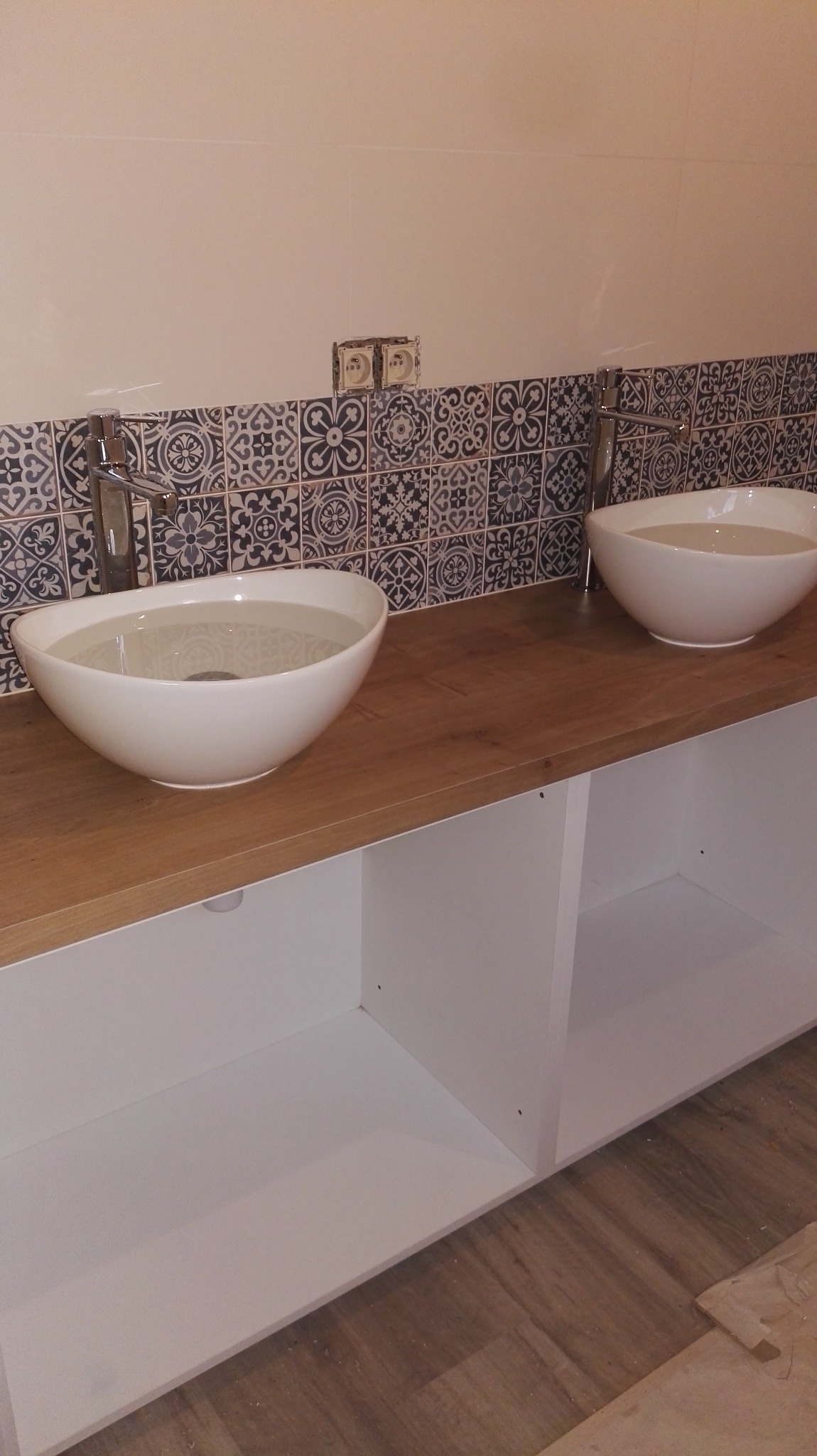 Dwie białe, owalne umywalki nablatowe z chromowanymi bateriami na drewnianym blacie. W tle dekoracyjne, wzorzyste płytki i białe ściany. Widoczne gniazdka elektryczne.