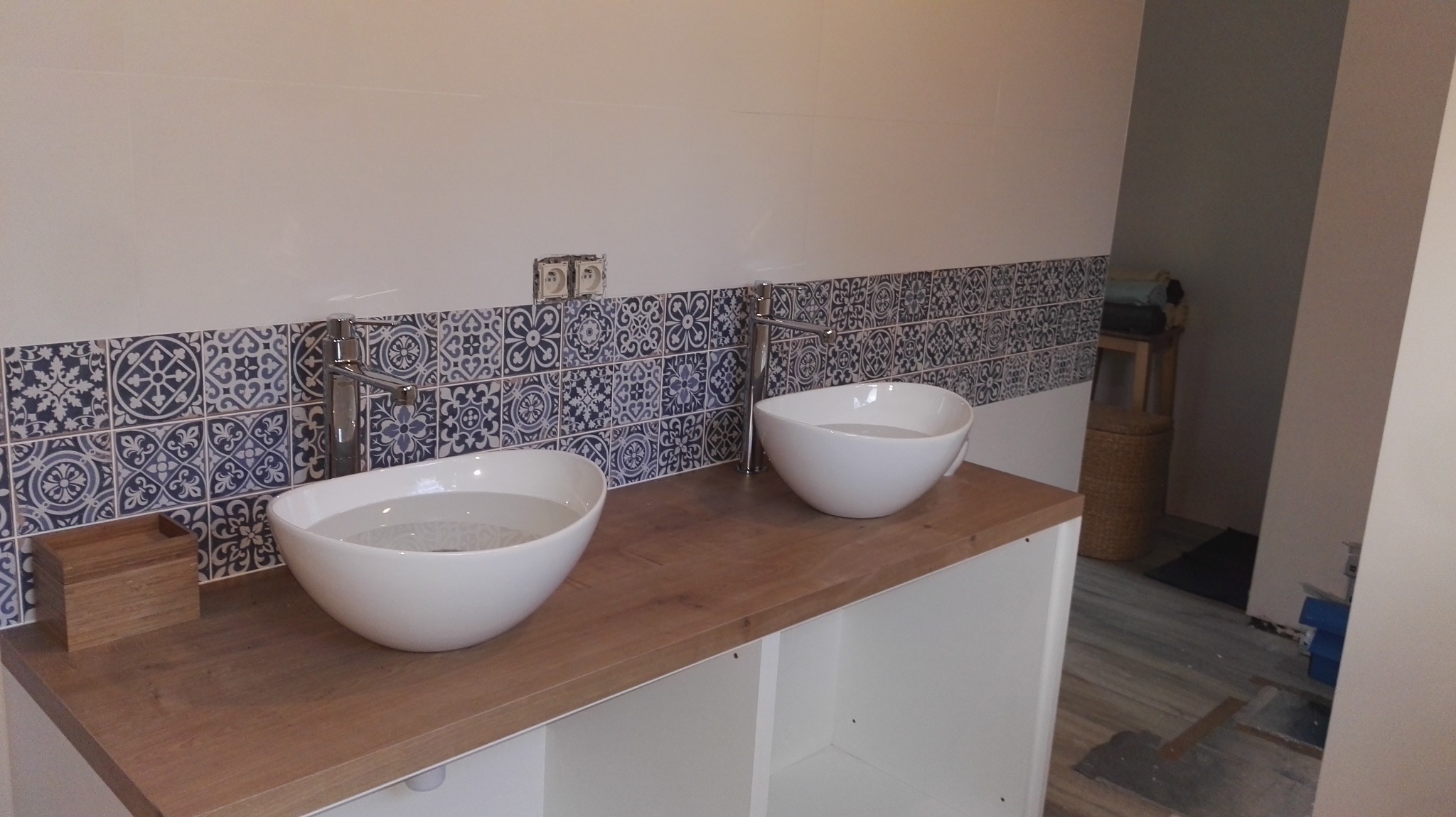 Dwie białe, okrągłe umywalki nablatowe z chromowanymi bateriami na drewnianym blacie. Nad nimi pas płytek w stylu marokańskim. Widoczne gniazdka elektryczne.
