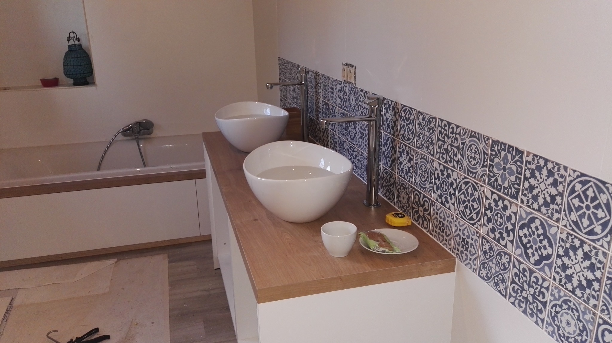 Wykończona łazienka z dwiema owalnymi umywalkami na drewnianym blacie, dekoracyjnymi płytkami i wanną w tle. Widoczne narzędzia glazurnicze.