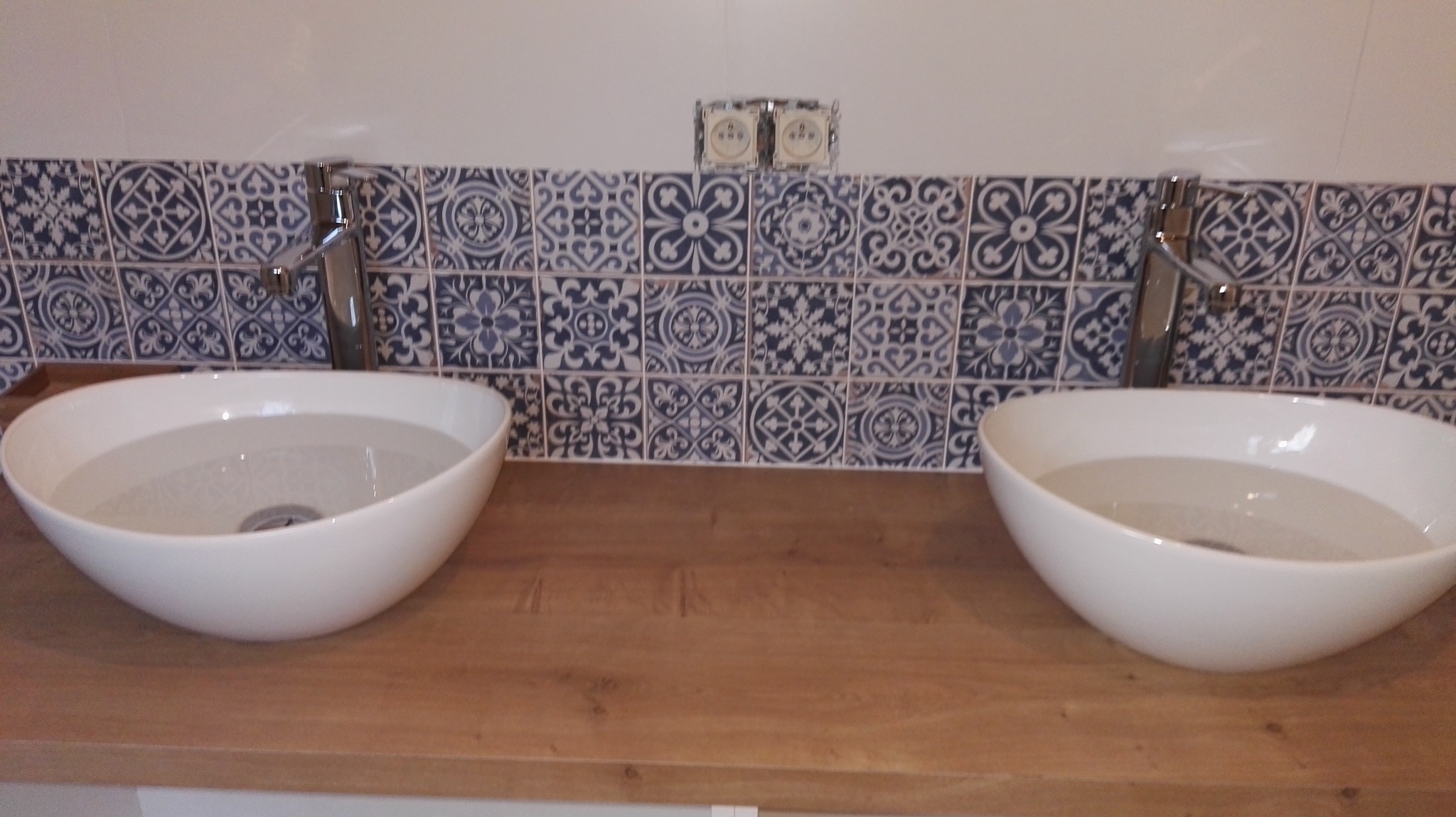 Dwie białe, owalne umywalki nablatowe z chromowanymi bateriami na drewnianym blacie. Nad nimi dekoracyjne, niebiesko-białe płytki ceramiczne i gniazdka elektryczne.