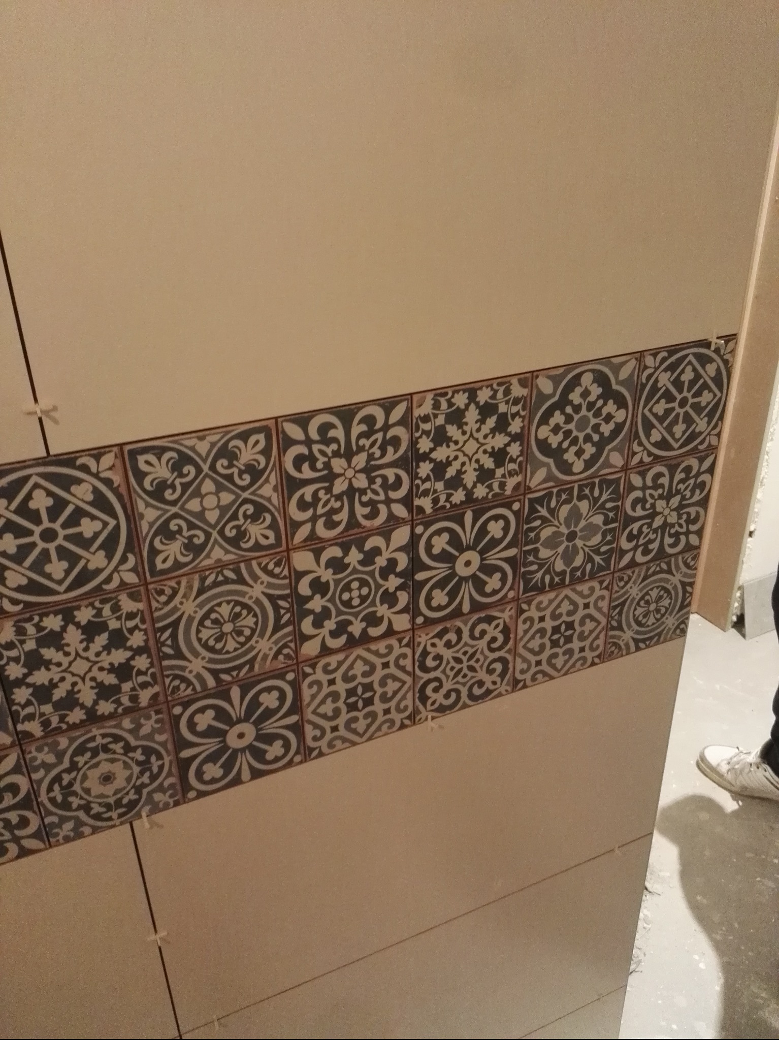 Ściana wyłożona beżowymi płytkami z dekoracyjnym pasem płytek w stylu vintage, z widocznymi krzyżykami dystansowymi, częściowo widoczna osoba w tle.