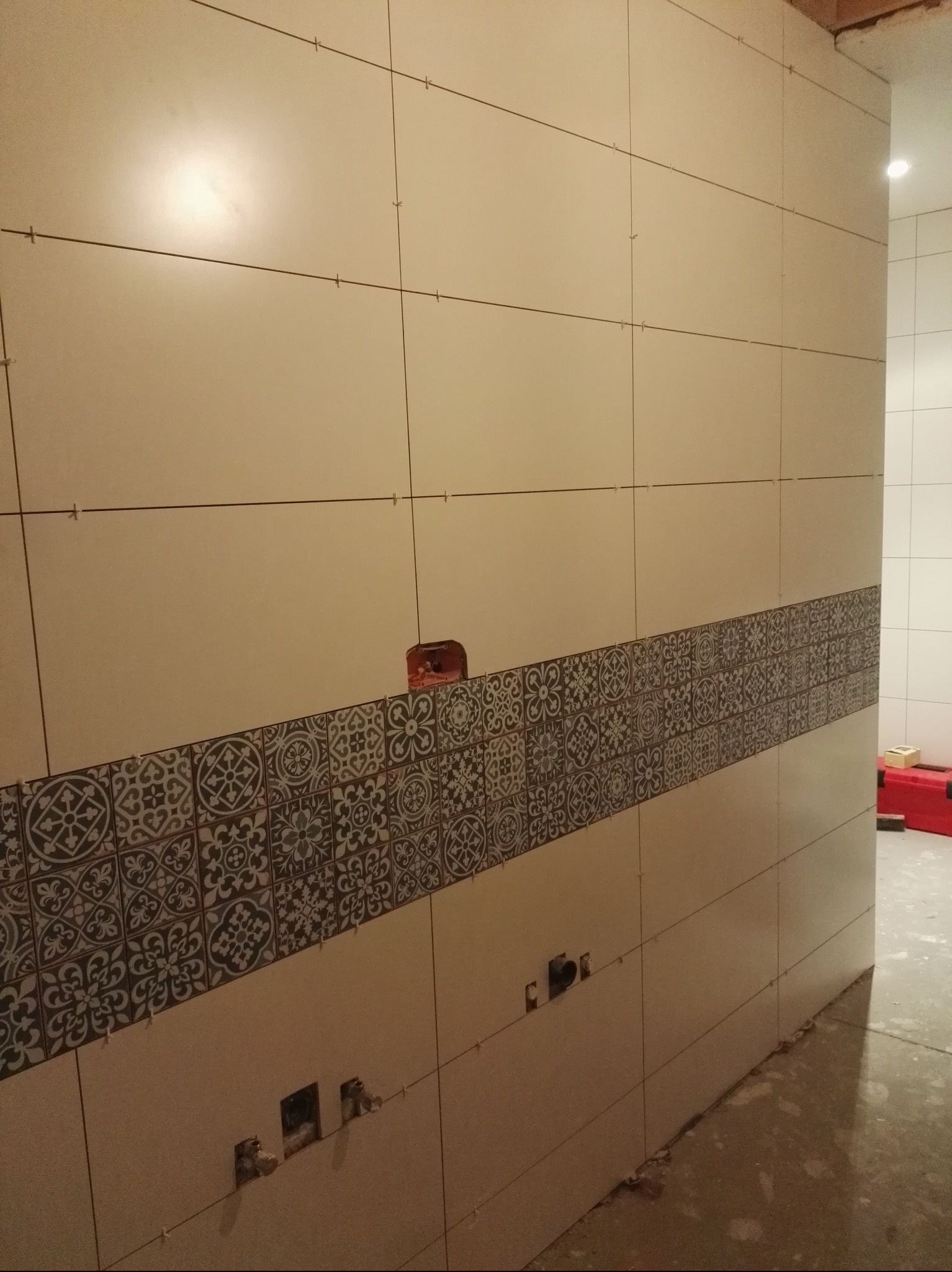 Ściana w trakcie układania płytek z dekoracyjnym pasem wzorzystych kafli. Widoczne krzyżyki dystansowe i otwory na instalacje. W tle fragment podłogi i narzędzia.