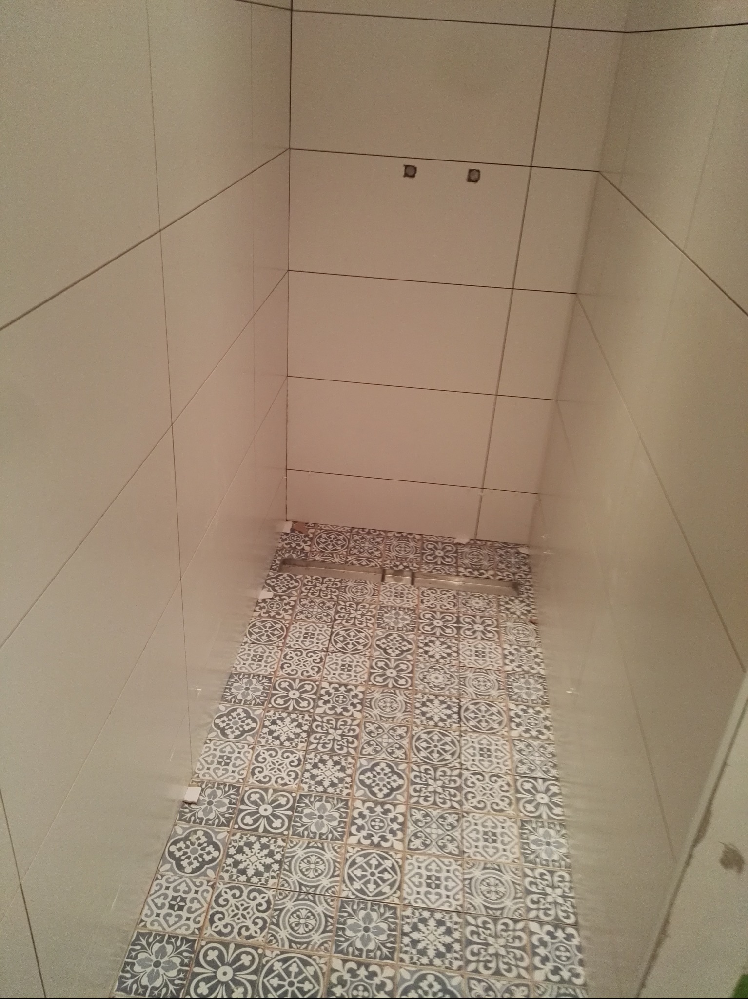 Wnętrze łazienki w trakcie prac wykończeniowych. Na ścianach jasne, gładkie płytki, na podłodze wzorzysta mozaika w odcieniach szarości i bieli. Widoczne niedokończone fragmenty podłogi.