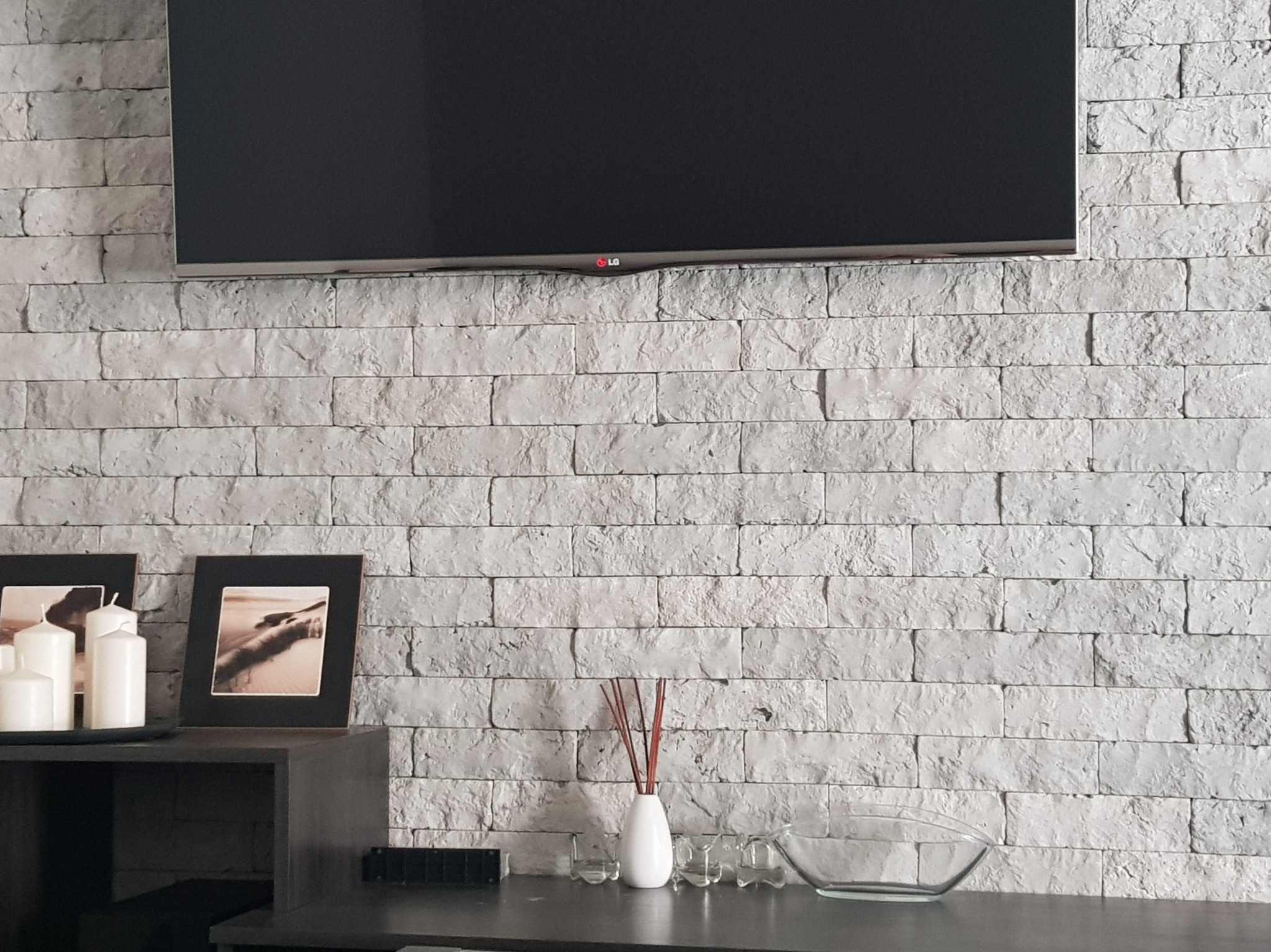Ściana z dekoracyjnej, szarej cegły z telewizorem LG. Na komodzie ramki ze zdjęciami, świece i dekoracje. Minimalistyczny wystrój wnętrza.