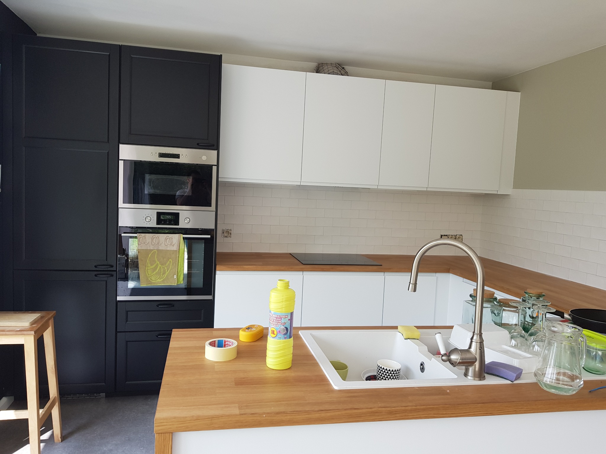 Nowoczesna kuchnia z czarno-białymi szafkami, drewnianym blatem i białym zlewem. Widoczne akcesoria kuchenne i środki czystości na blacie. Minimalistyczny design.