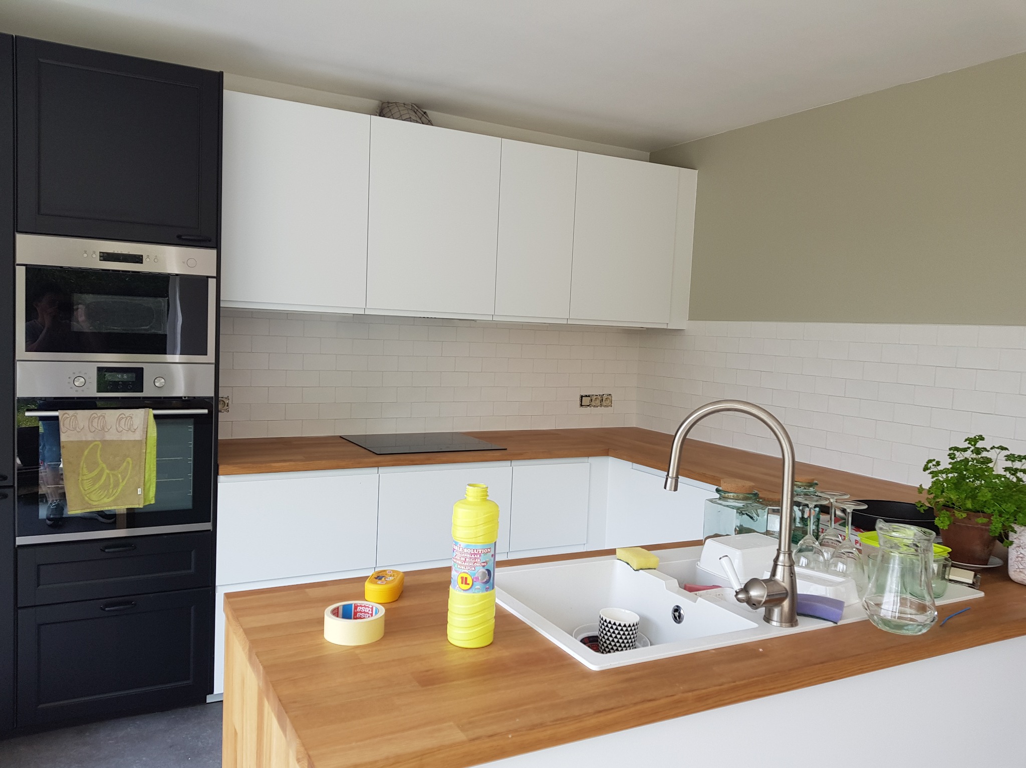 Nowoczesna kuchnia z białymi szafkami, drewnianym blatem i czarną zabudową piekarnika. Na blacie akcesoria kuchenne i środki czystości. Białe płytki na ścianie.