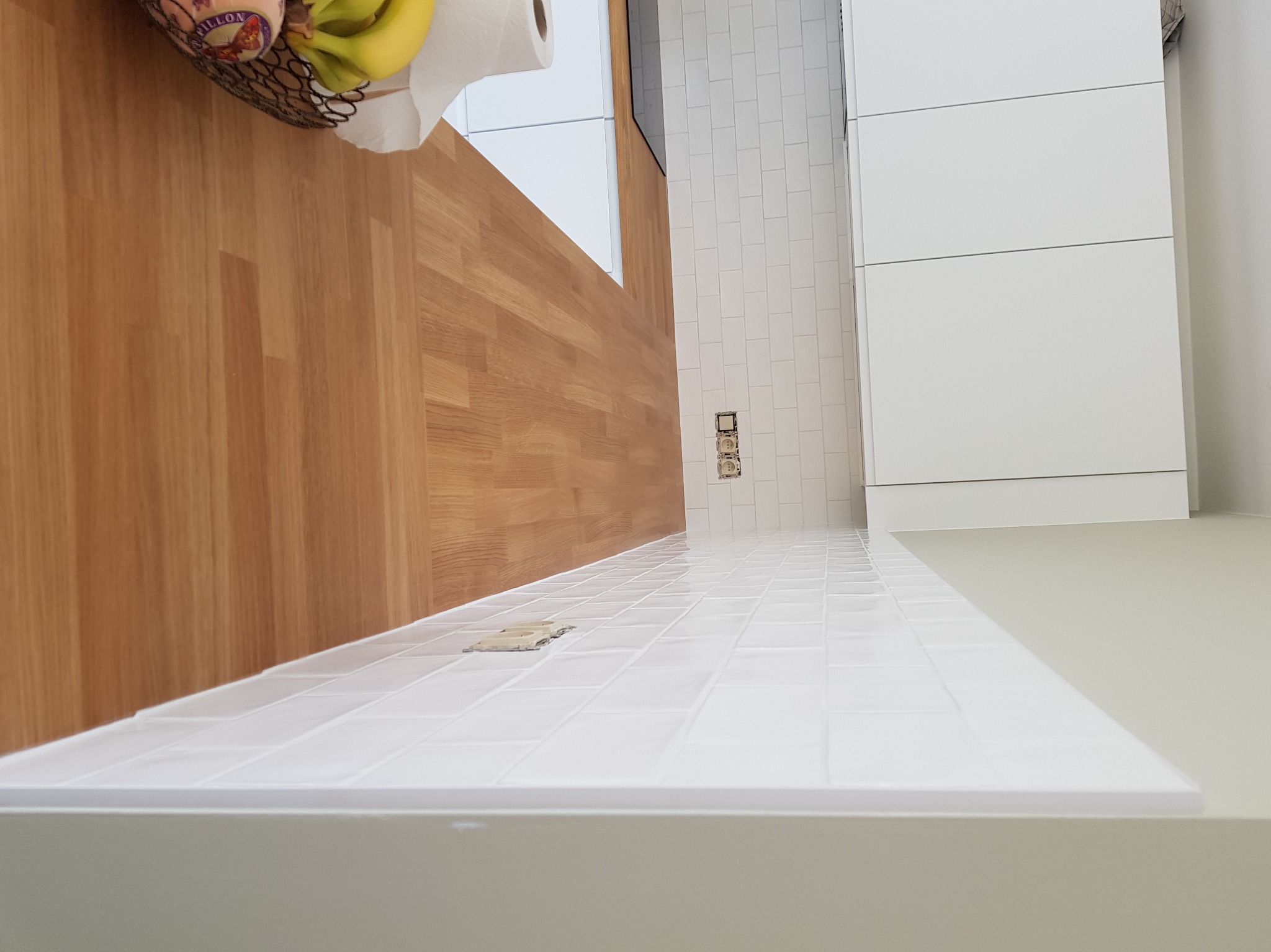 Białe, prostokątne płytki ułożone na ścianie w kuchni, widoczne gniazdka elektryczne. Na blacie koszyk z owocami i rolka papieru. Drewniana okładzina ściany.