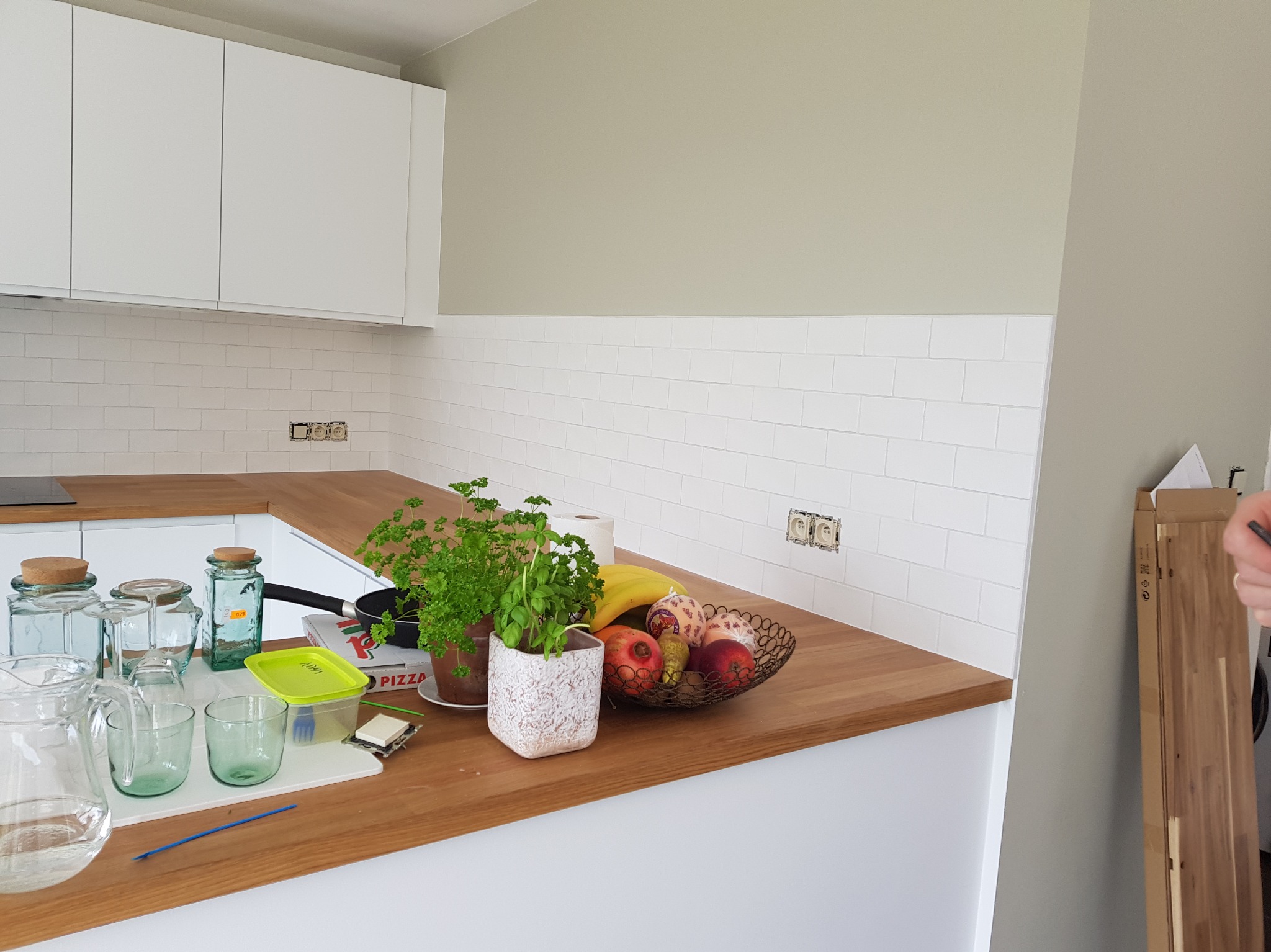 Jasna kuchnia w trakcie wykończenia: białe płytki na ścianie, drewniany blat, gniazdka elektryczne bez osprzętu, świeże zioła i owoce na blacie.
