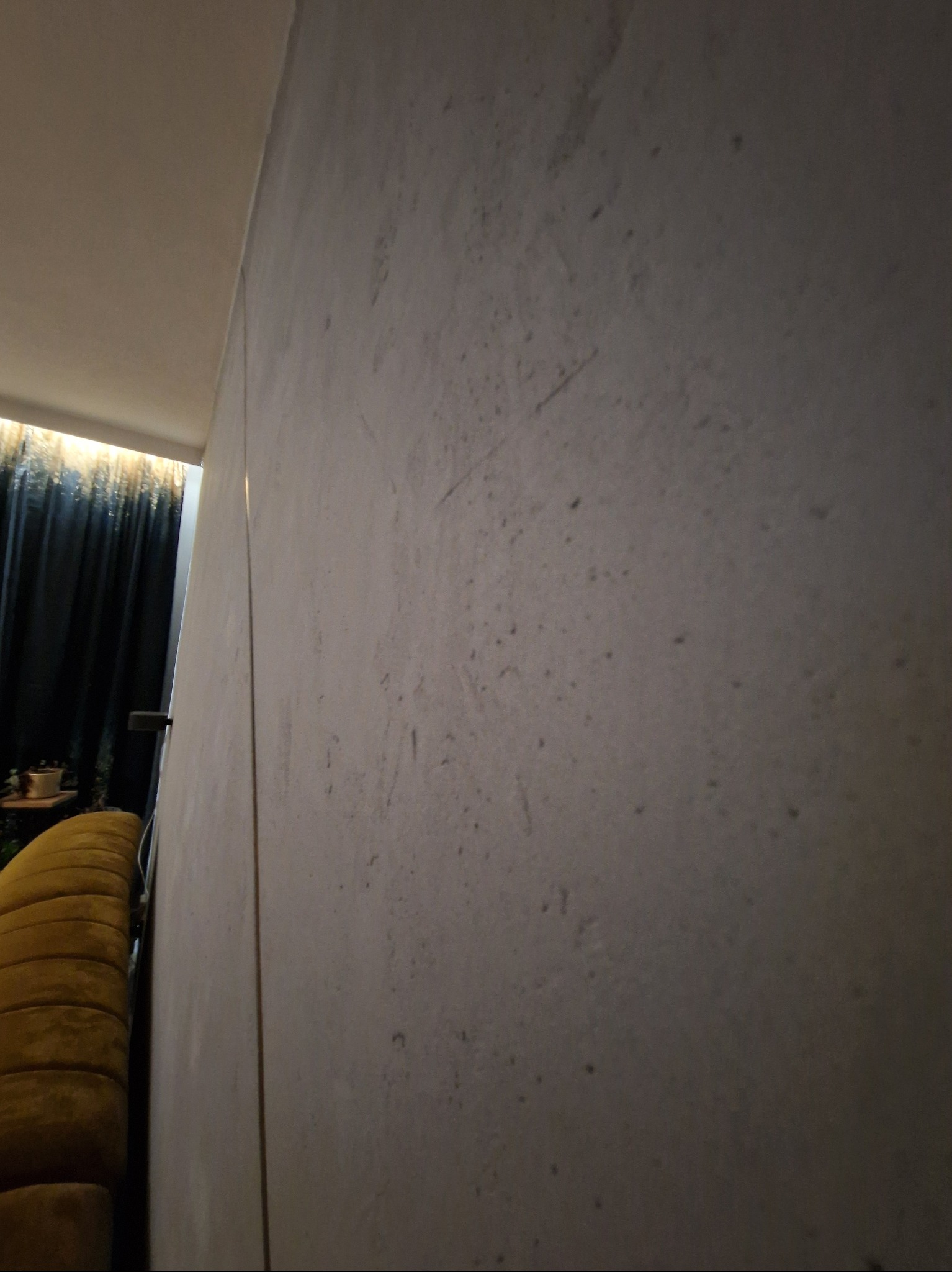 Ściana z efektem betonu w szarościach, widoczna z boku, z fragmentem sofy w odcieniach złota i zasłonami w tle. Minimalistyczne wykończenie wnętrza.