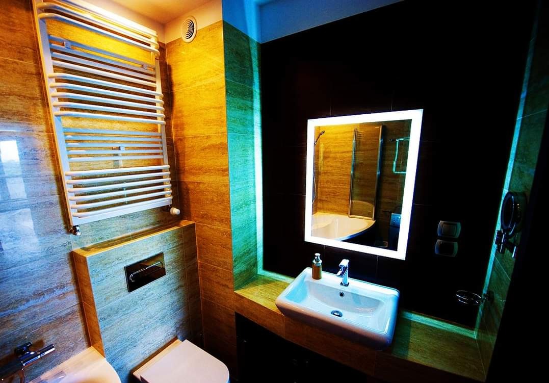 Nowoczesna łazienka z podświetlanym lustrem, ceramicznymi płytkami imitującymi drewno, białym grzejnikiem drabinkowym i umywalką nablatową z chromowaną baterią.