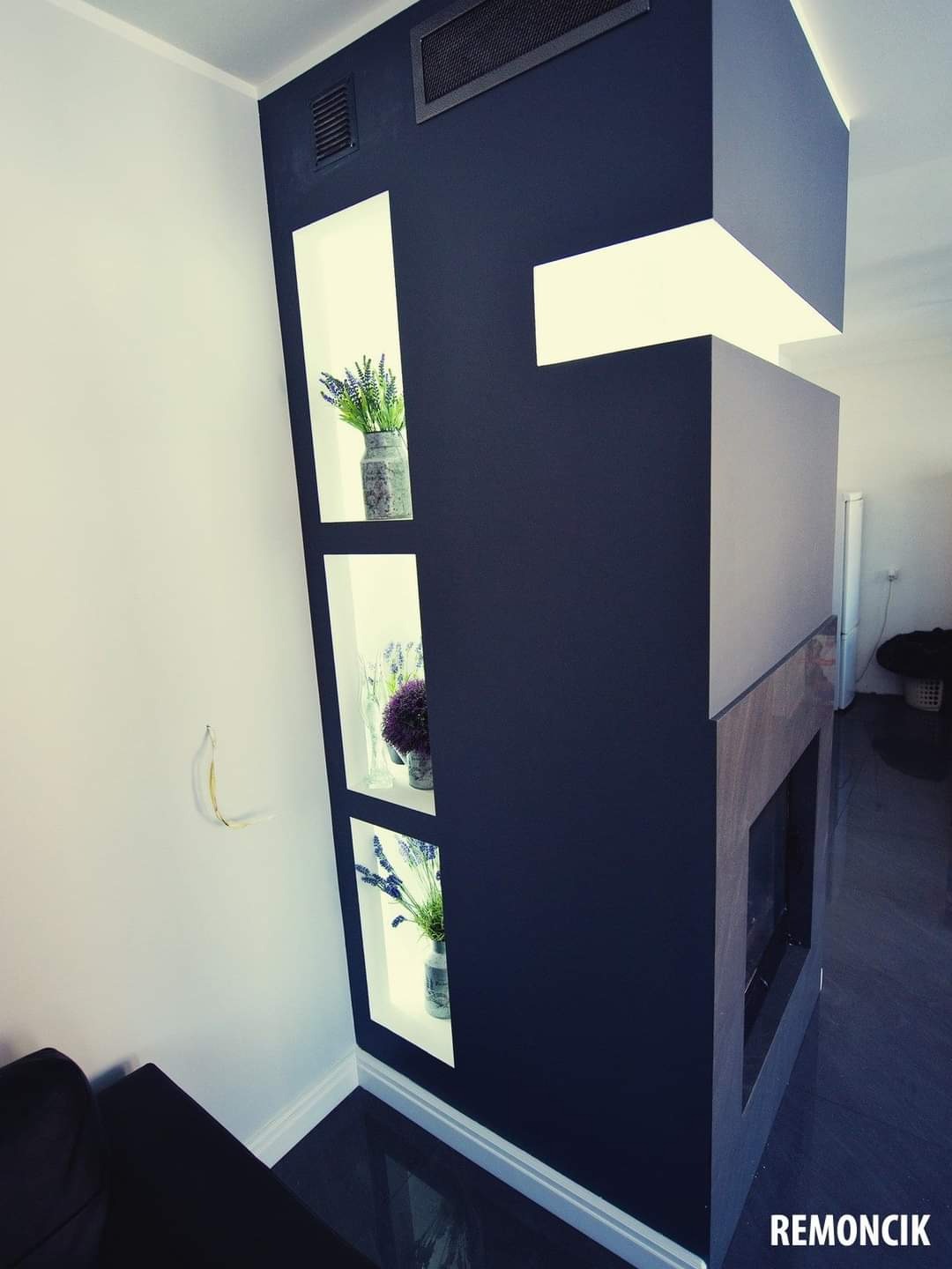 Nowoczesna ściana działowa w kolorze granatowym z wnękami podświetlonymi światłem, w których umieszczono szklane wazony z lawendą i innymi kwiatami, obok widoczny fragment obudowy kominka z szarą...