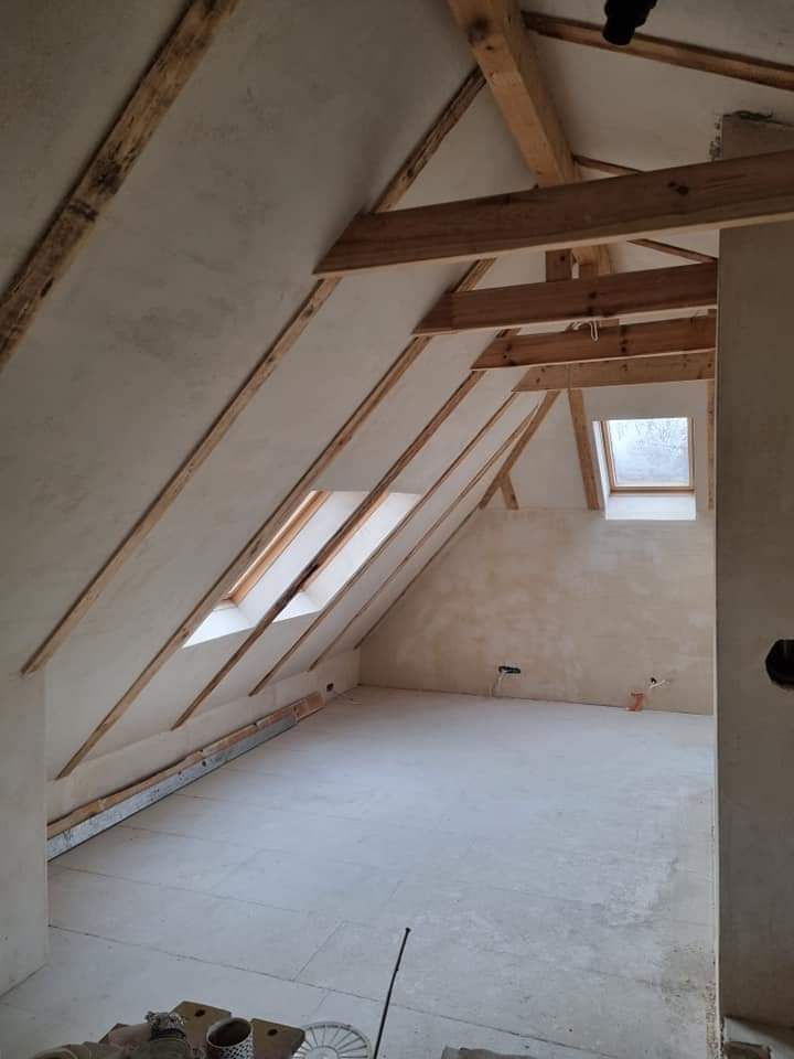 Poddasze w stanie surowym: otynkowane ściany, drewniana konstrukcja dachu z oknami połaciowymi, betonowa wylewka na podłodze, widok z dołu na górę przez otwarte drzwi.