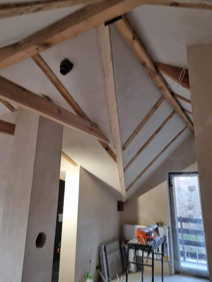 Wnętrze poddasza w trakcie prac wykończeniowych: surowe drewniane belki stropowe, ściany przygotowane pod malowanie z widocznym otworem, okno balkonowe i narzędzia na stole roboczym.