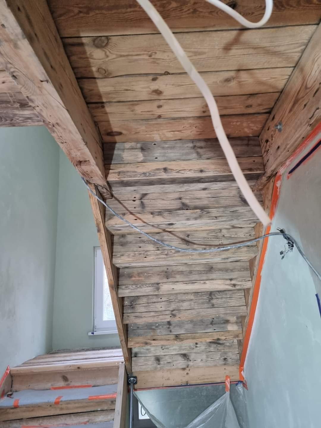 Surowe drewniane schody w trakcie budowy, zabezpieczone taśmą malarską, z widocznymi przewodami elektrycznymi i surowym wykończeniem ścian.