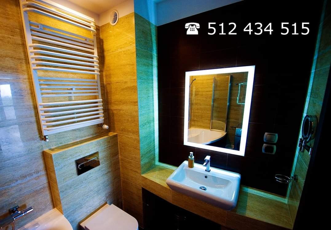 Nowoczesna łazienka z oświetlonym lustrem, grzejnikiem drabinkowym i elementami wykończenia w odcieniach beżu i brązu, uzupełniona o numer telefonu.