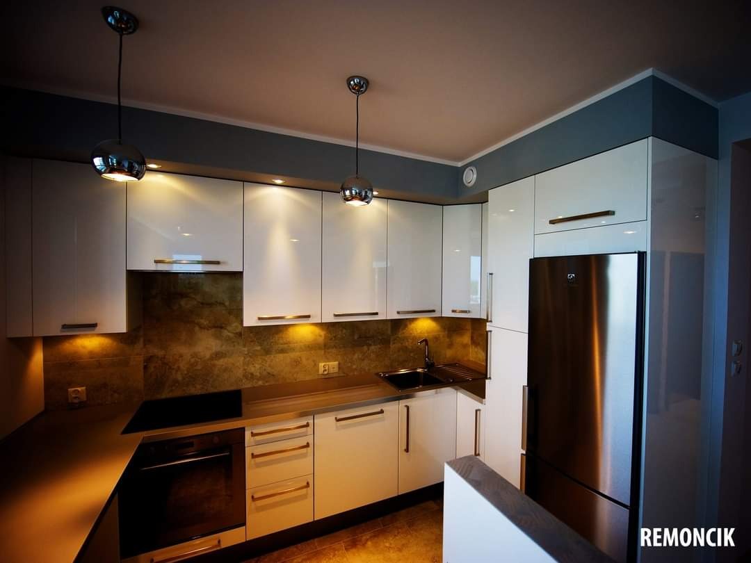 Nowoczesna, lśniąca kuchnia z białymi szafkami, złotymi uchwytami i marmurkowym backsplash'em, oświetlona wiszącymi lampami w stylu glamour i podświetleniem LED pod szafkami.