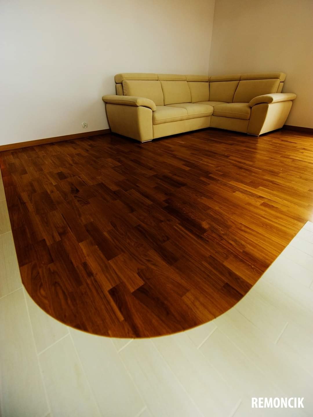 Salon z nową, ciemnobrązową podłogą parkietową, przechodzącą w jasne płytki, oraz beżową sofą narożną przy białej ścianie.