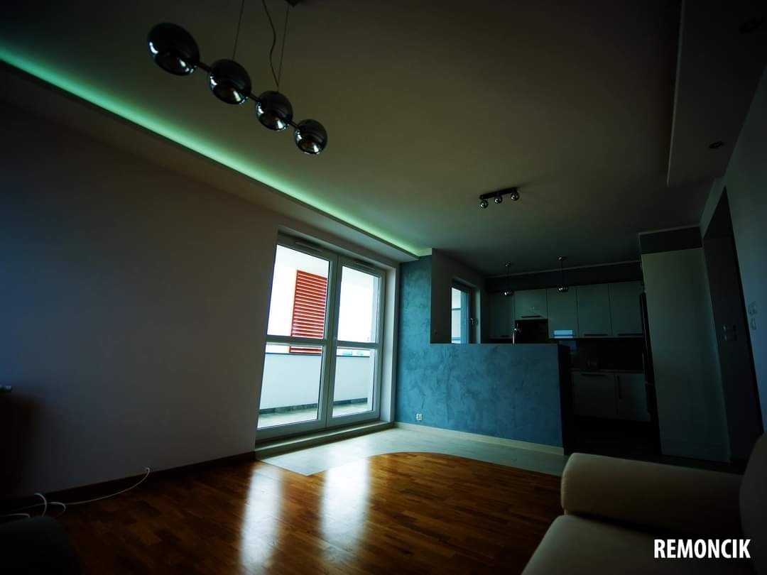 Nowoczesne wnętrze mieszkania po remoncie, z parkietem, gładzią na ścianie w odcieniach szarości i niebieskiego, oświetleniem LED pod sufitem i widokiem na zabudowany balkon z żaluzjami.