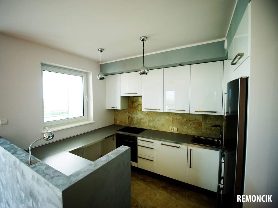 Nowoczesna kuchnia z białymi, lakierowanymi szafkami, szarym blatem i mozaiką na ścianie, oświetlona dwoma chromowanymi lampami wiszącymi i światłem dziennym z okna.