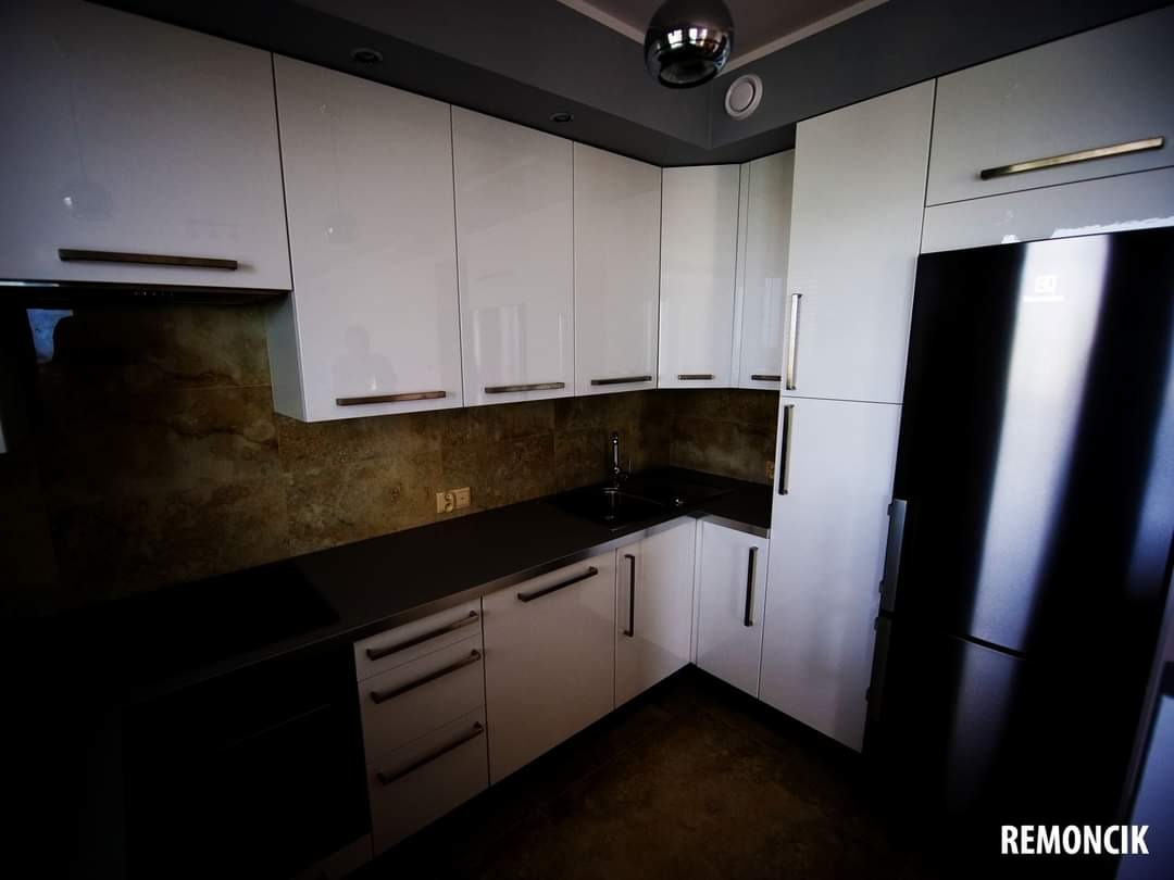 Nowoczesna, lśniąca kuchnia z białymi szafkami, ciemnym blatem i brązowymi płytkami na ścianie, uzupełniona o czarną lodówkę z metalowymi uchwytami.