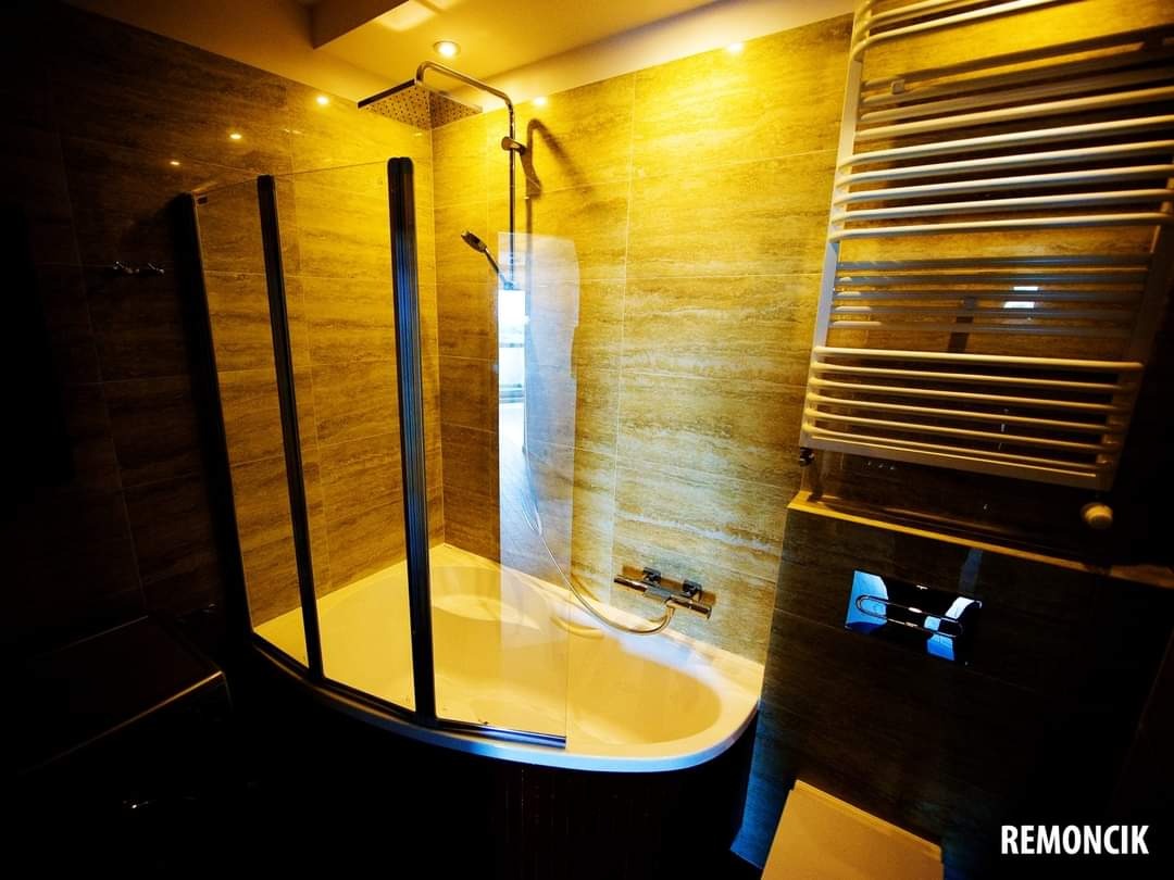 Nowoczesna łazienka z kabiną prysznicową typu walk-in, grzejnikiem drabinkowym i płytkami imitującymi kamień w ciepłych odcieniach, oświetlona punktowymi lampami sufitowymi.