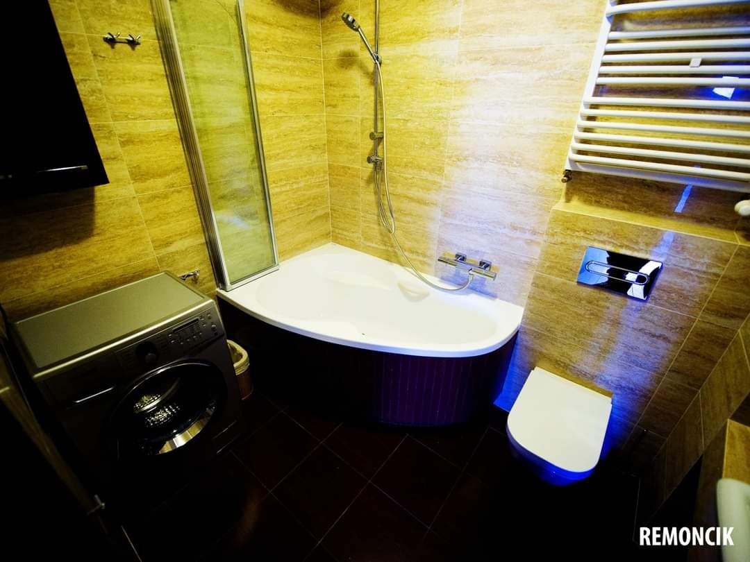 Ukończona łazienka z beżowymi płytkami imitującymi trawertyn, narożną wanną z ciemnoczerwoną obudową, pralką obok kabiny prysznicowej i grzejnikiem drabinkowym. Uwagę zwraca podświetlenie LED...