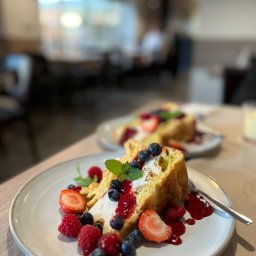 DAMIAN LEGAT - Delikatny deser: kawałek ciasta z kremem, świeżymi truskawkami, malinami i borówkami, polany czerwonym sosem i udekorowany listkiem mięty, na jasnym talerzu z widelcem, w tle rozmyte wnętrze kawiarni.