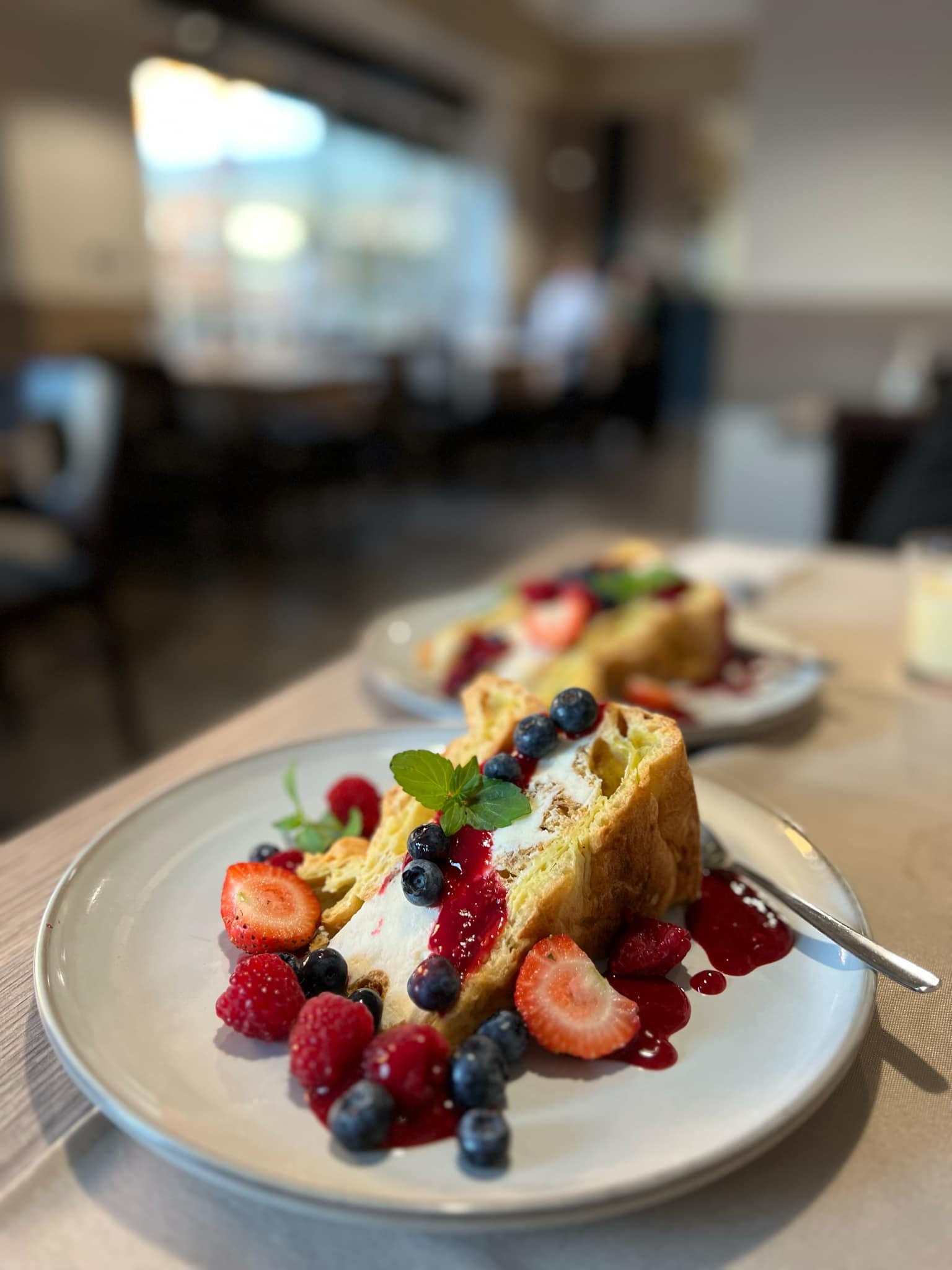 Delikatny deser: kawałek ciasta z kremem, świeżymi truskawkami, malinami i borówkami, polany czerwonym sosem i udekorowany listkiem mięty, na jasnym talerzu z widelcem, w tle rozmyte wnętrze kawiarni.