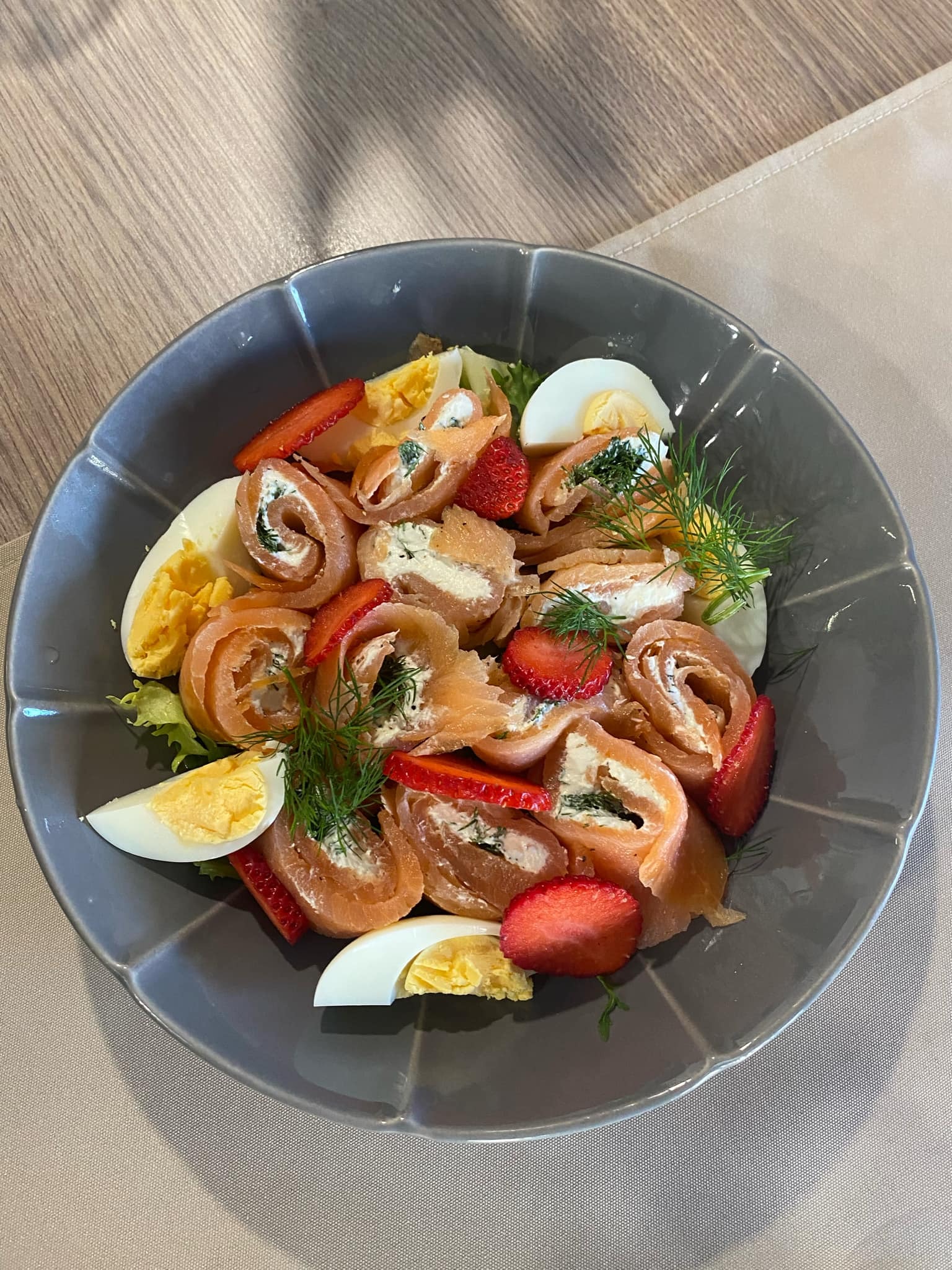 Szary talerz z roladkami z łososia nadziewanymi kremowym serem i koperkiem, udekorowanymi ćwiartkami truskawek, jajkiem na twardo i koperkiem.