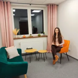 Kobieta z długimi włosami siedzi na pomarańczowym krześle w gabinecie z zieloną kanapą i małymi stolikami, trzymając clipboard.