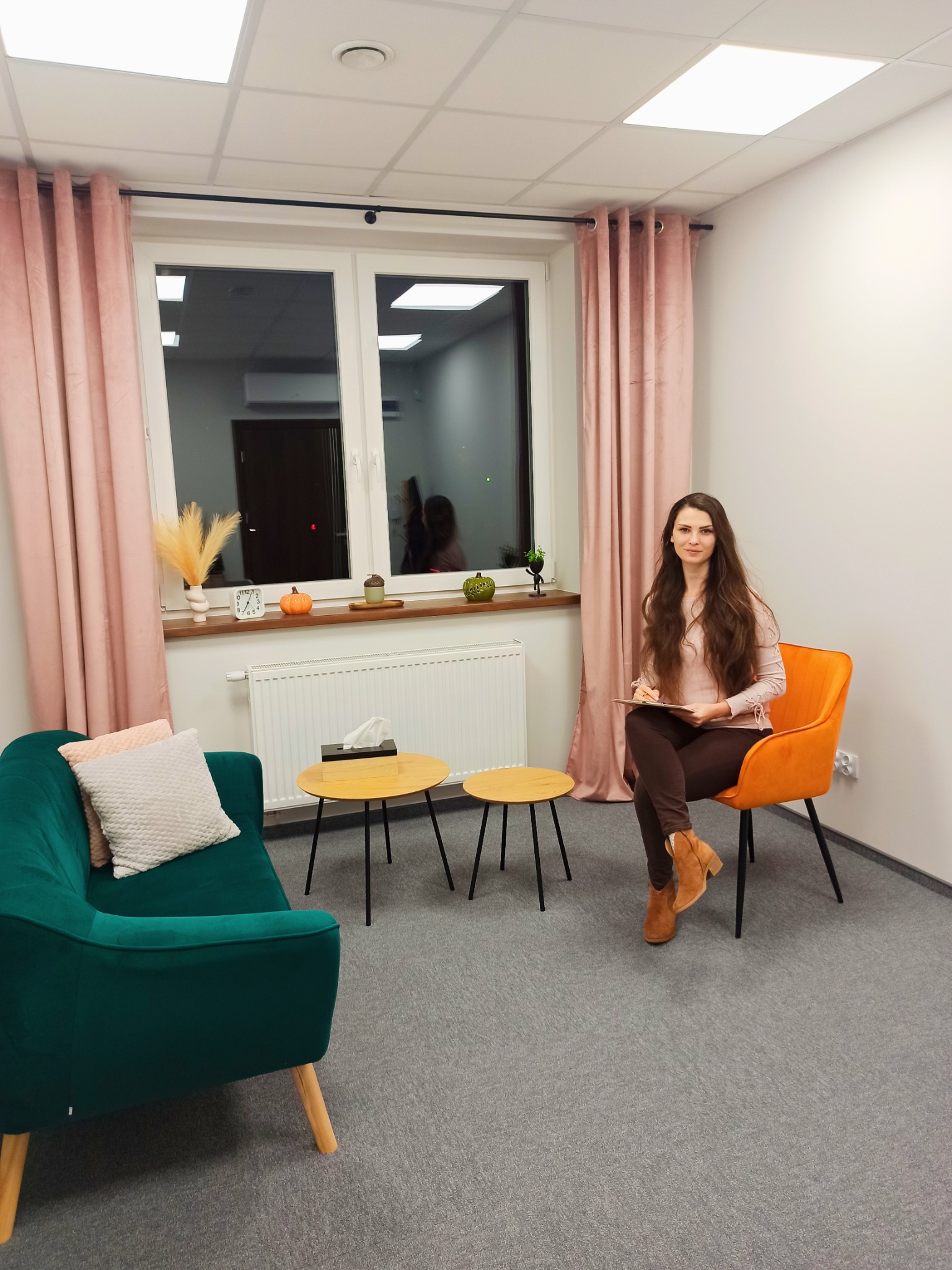 Kobieta z długimi włosami siedzi na pomarańczowym krześle w gabinecie z zieloną kanapą i małymi stolikami, trzymając clipboard.