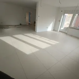 Jasne, duże płytki podłogowe ułożone w nowym mieszkaniu, widoczne krzyżyki dystansowe, przygotowane do fugowania, ściany pomalowane na biało, widoczne kable elektryczne i okna z widokiem na budynek...