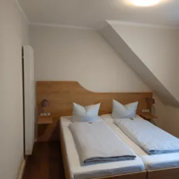 Pokój z dwoma pojedynczymi łóżkami z drewnianymi ramami i zagłówkiem, szafkami nocnymi z lampkami, poddasze z oknem dachowym, białe ściany i brązowa podłoga.