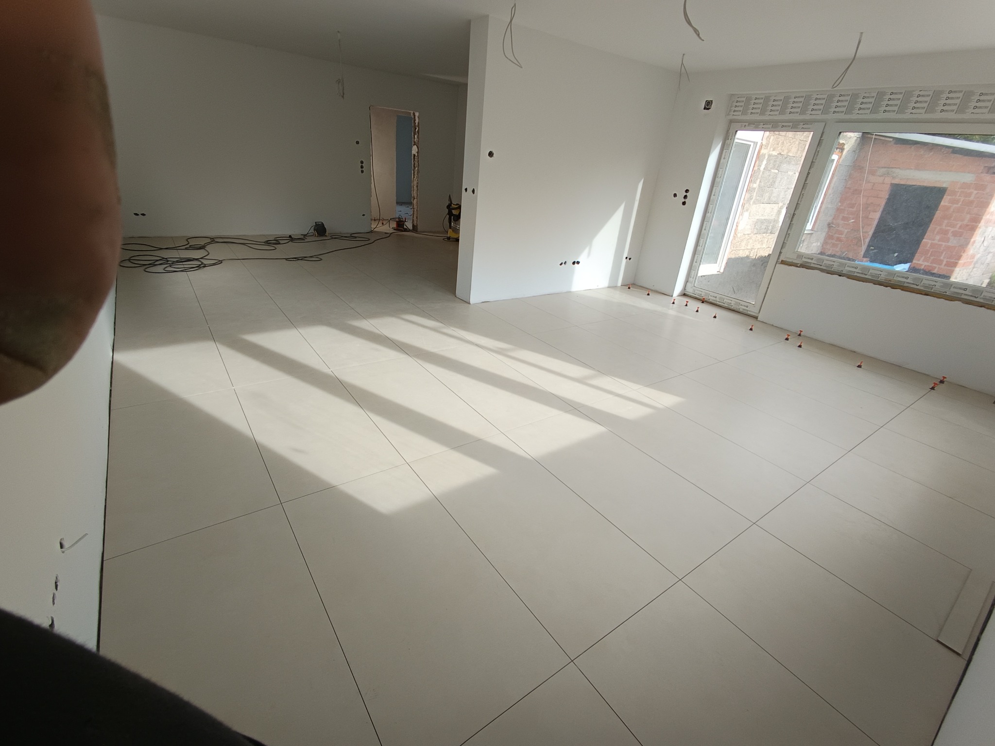 Jasne, duże płytki podłogowe ułożone w nowym mieszkaniu, widoczne krzyżyki dystansowe, przygotowane do fugowania, ściany pomalowane na biało, widoczne kable elektryczne i okna z widokiem na budynek...