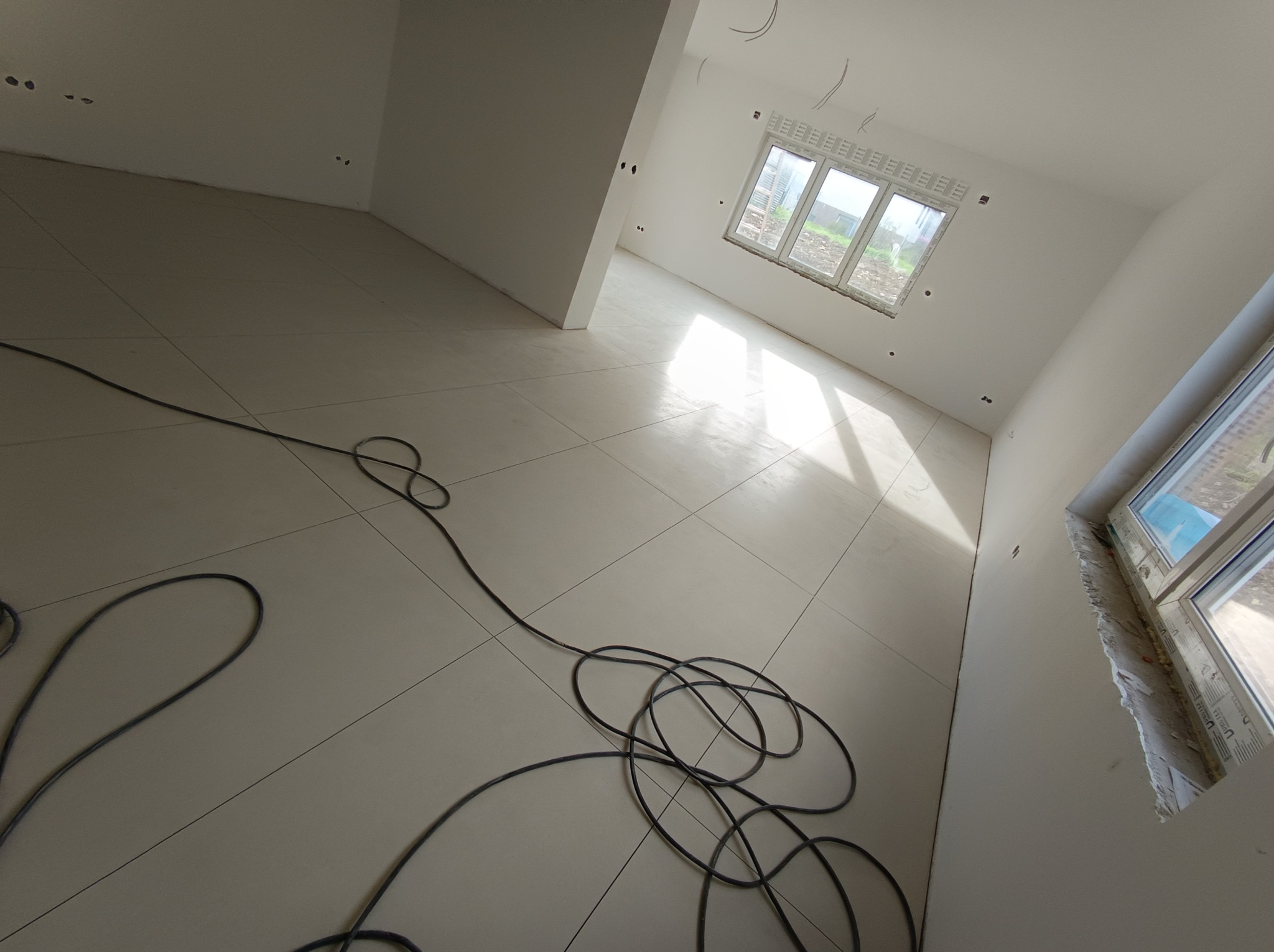 Wnętrze w stanie surowym: białe ściany, jasna kafelkowana podłoga, okna z widokiem na zewnątrz, zwisające kable elektryczne na podłodze.