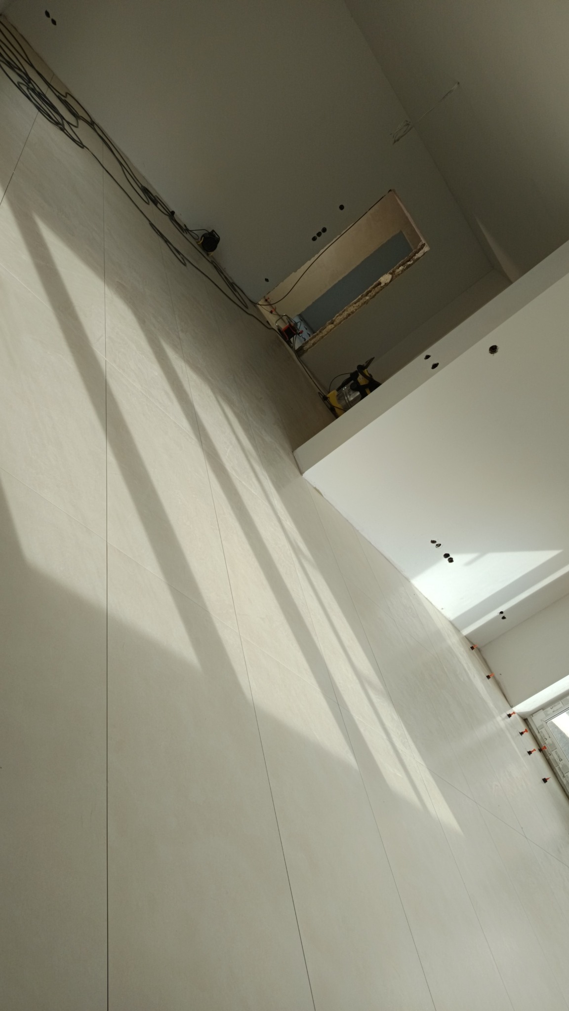 Wnętrze w trakcie wykańczania: Ściana wyłożona jasnymi, pionowymi płytkami, widoczne przewody elektryczne na suficie, otwór w suficie z widocznym oświetleniem i narzędziami, oraz elementy...