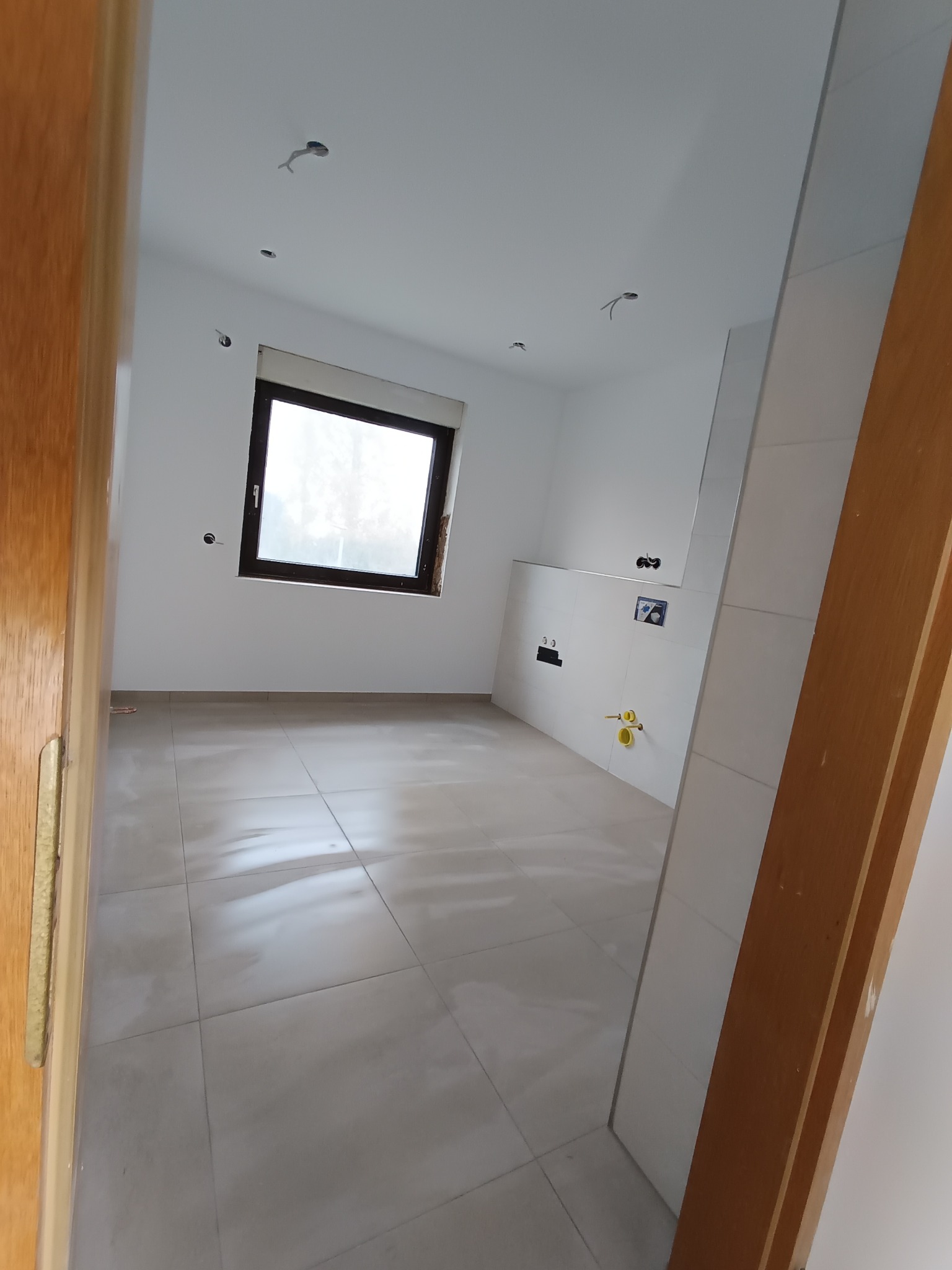 Wnętrze remontowanej łazienki z nowymi płytkami podłogowymi, przygotowanymi instalacjami hydraulicznymi i elektrycznymi, widoczne otwory w suficie na oświetlenie, okno z ciemną ramą i fragment...