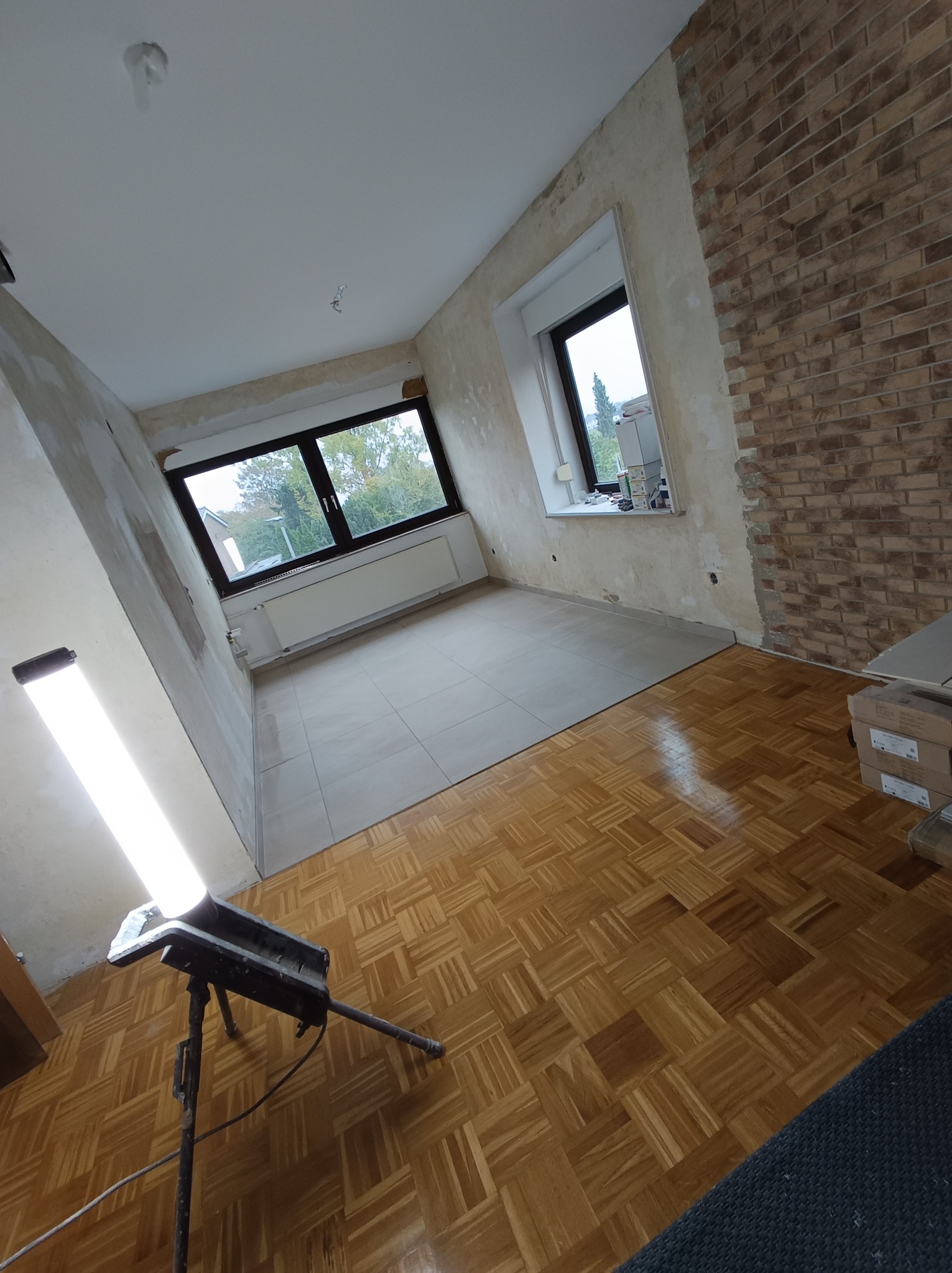 Pokój w trakcie remontu: częściowo położone płytki podłogowe, fragment ściany z cegły, surowe ściany przygotowane do malowania, widoczna lampa robocza.